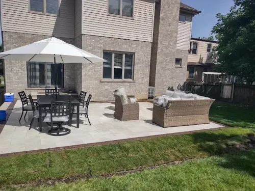 A patio with a dining set under an umbrella, wicker armchairs, and a sofa behind a house with light brick siding.