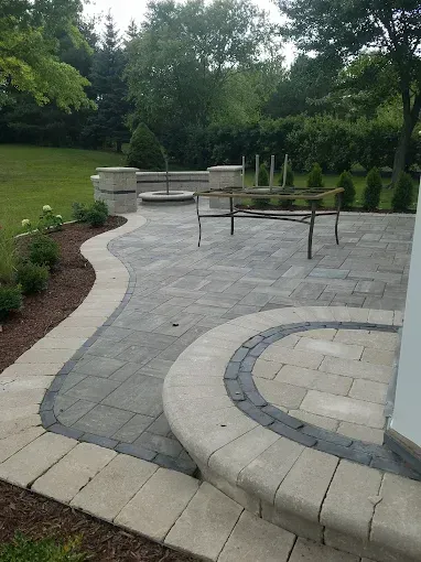 A backyard stone paver patio featuring a curved retaining wall bench, a fire pit, and a metal table frame.