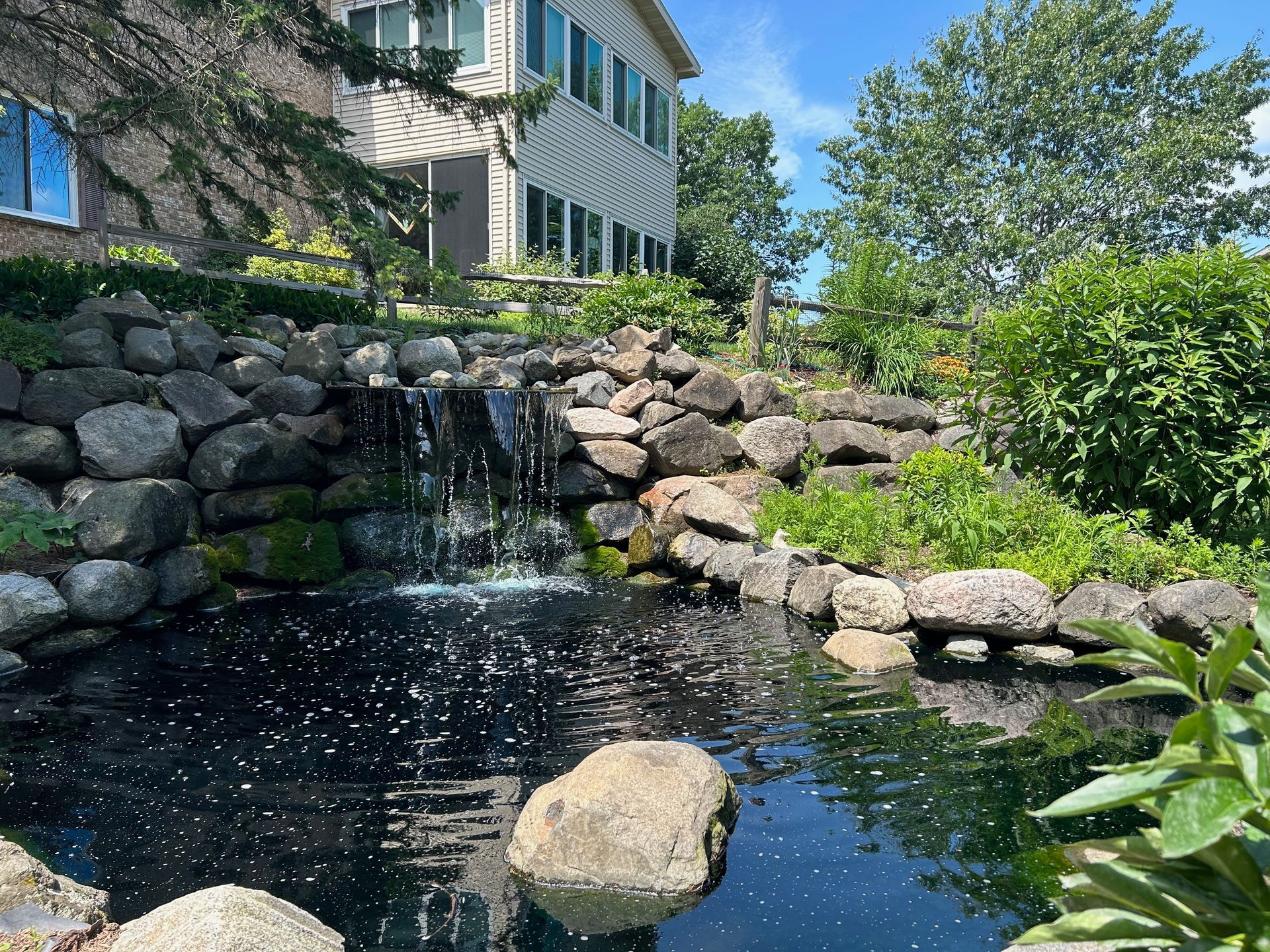 There is a waterfall in the middle of a pond in front of a house.