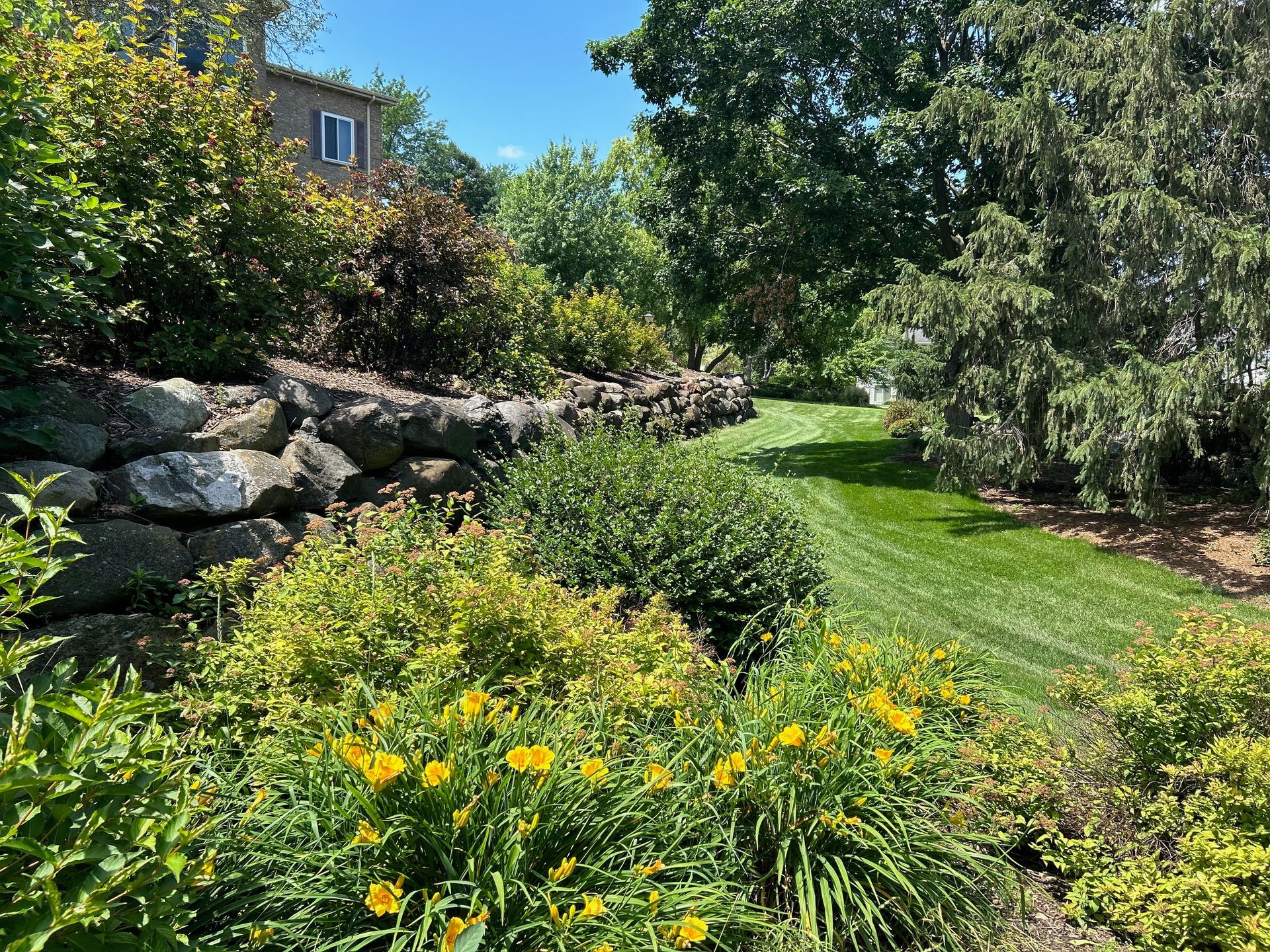 A lush green lawn surrounded by trees and flowers with a stone wall in the background.