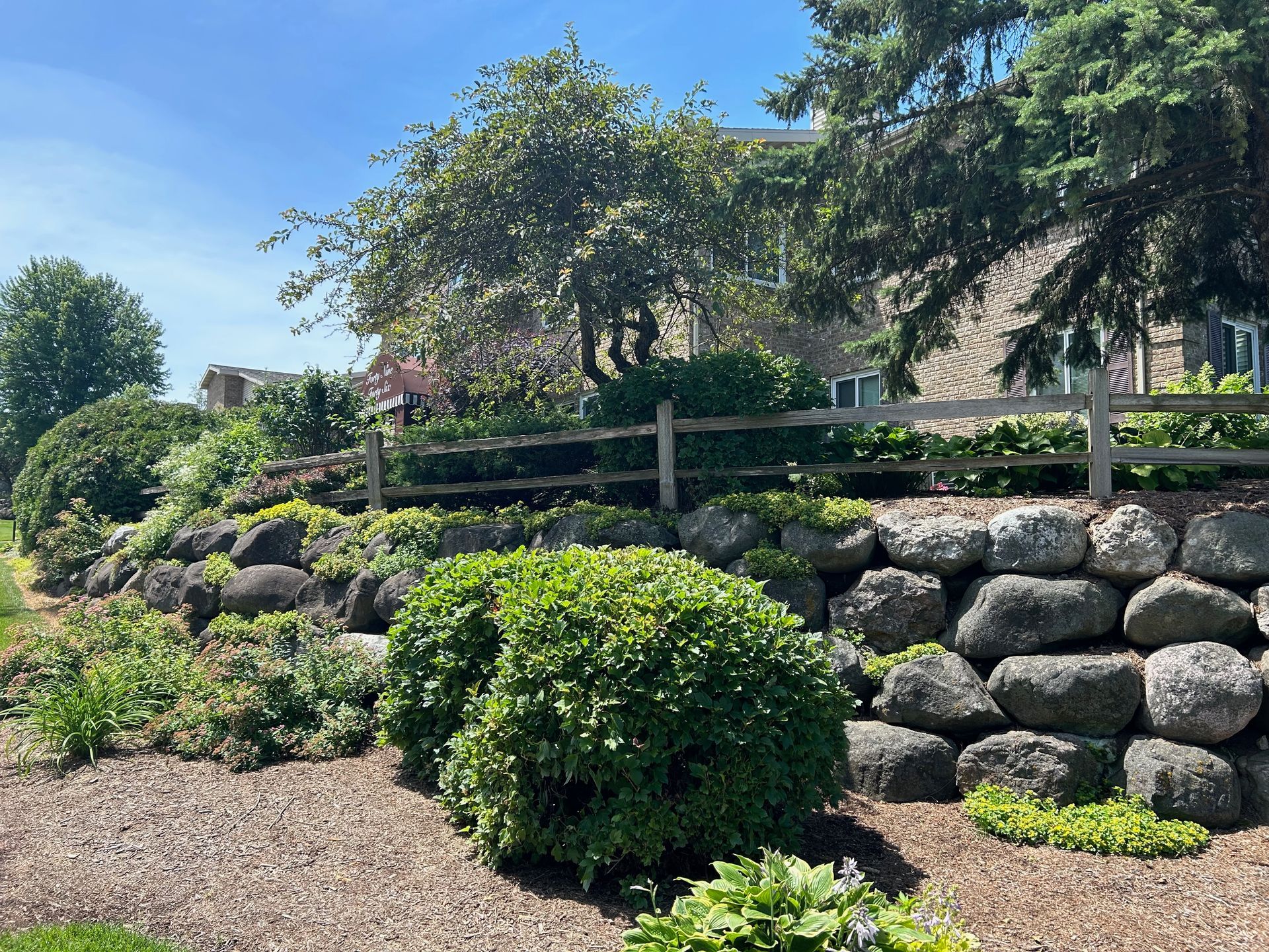 A stone wall with a wooden fence and bushes in front of a house.