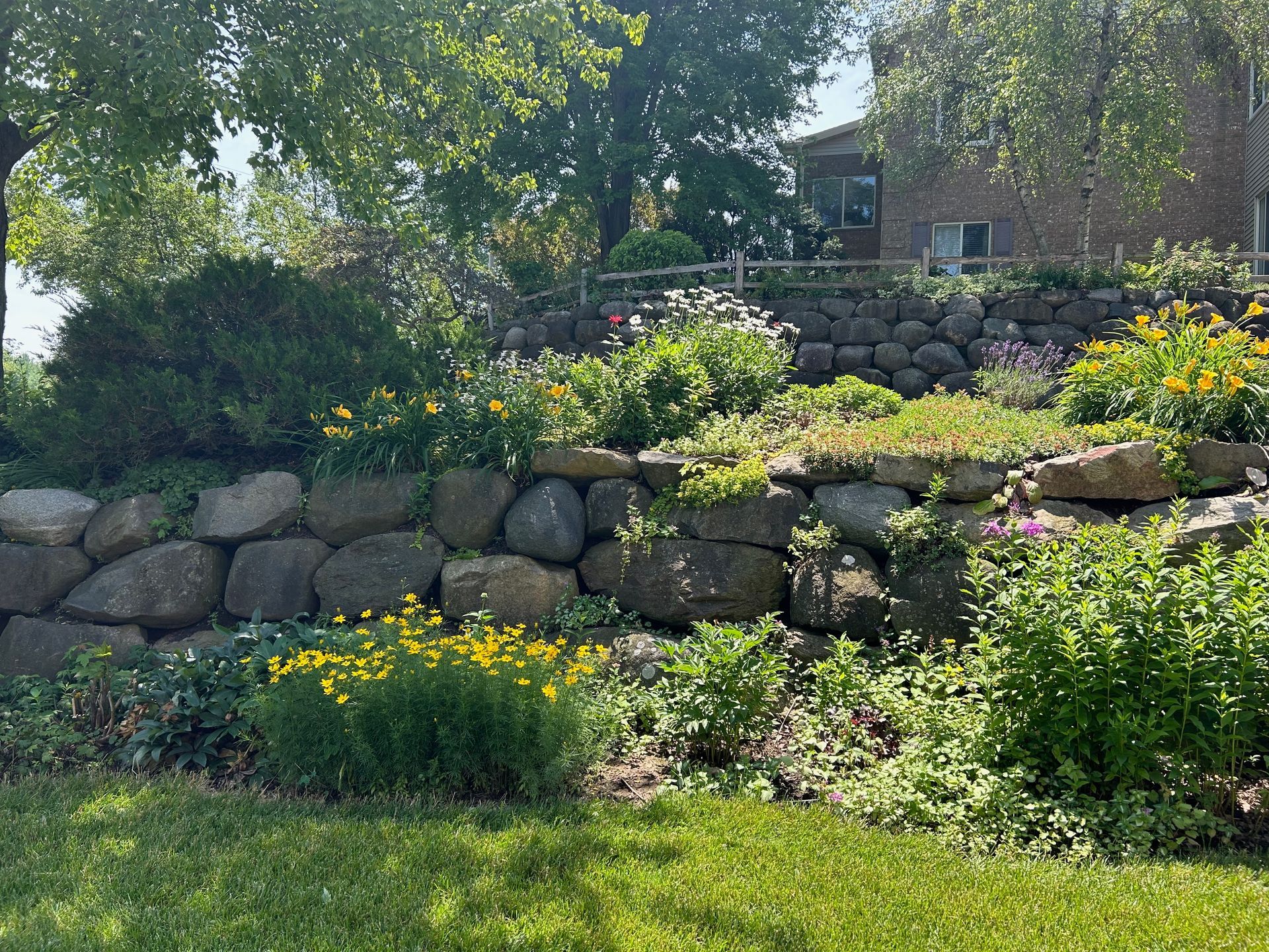 A stone wall surrounded by flowers and shrubs in a garden with a house in the background.