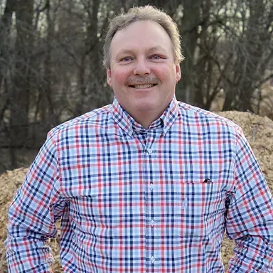 A man in a plaid shirt is standing in front of a pile of hay.