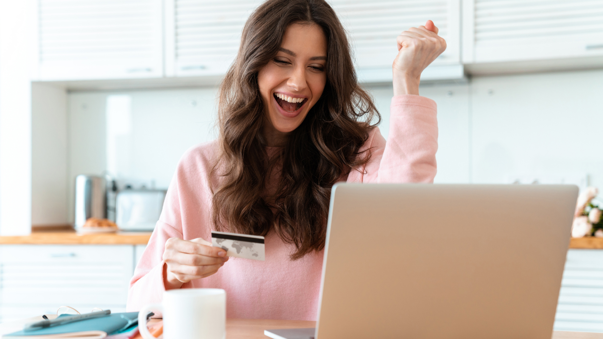 Woman with brown hair cheers while holding a credit card and looking at a laptop in a kitchen.