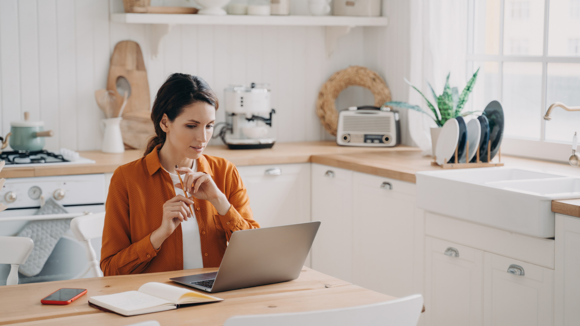 Woman in orange shirt working on a laptop at a kitchen table, looking focused. Notebook and phone are also on the table.