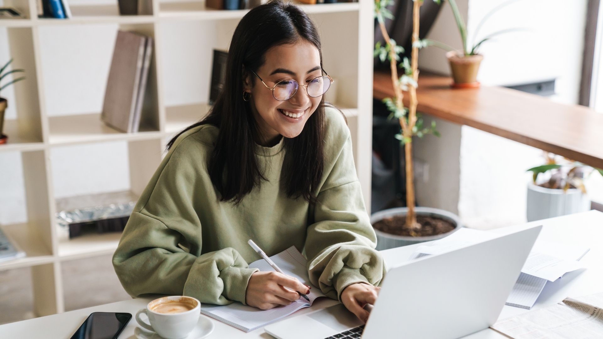 Young woman with glasses smiles while working on a laptop at a table with a coffee and notebook.