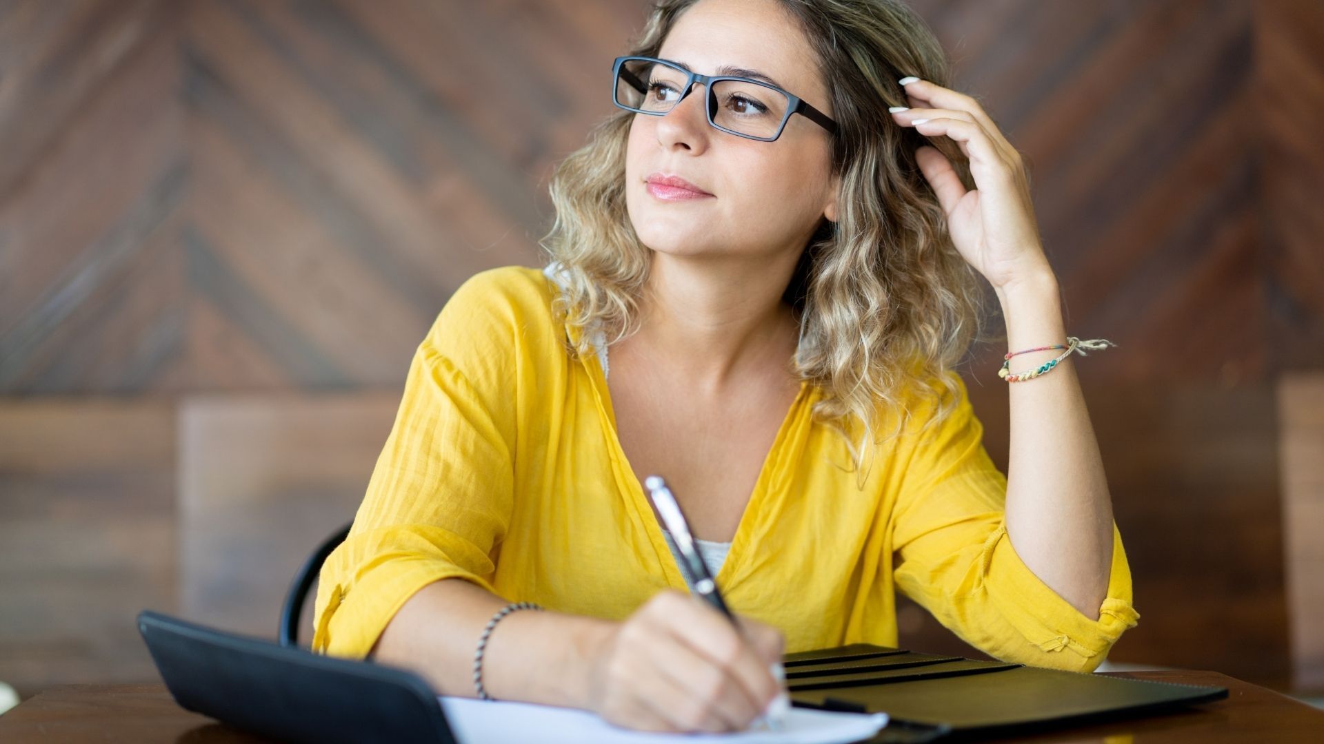 Woman in yellow top and glasses, writing at a desk, looking thoughtful, in front of a wood paneled wall.
