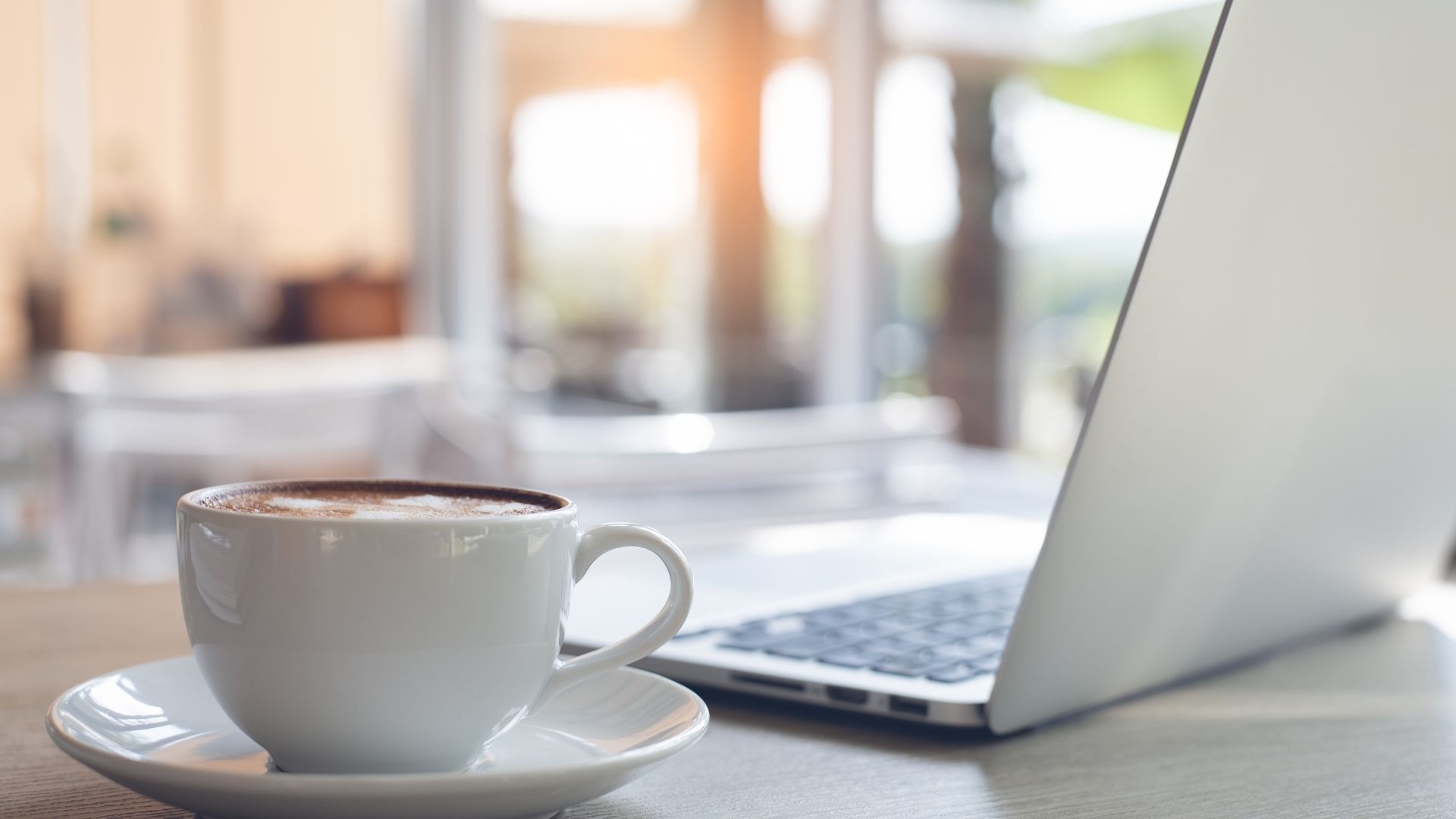 Cup of coffee with a laptop on a table, near a bright window.