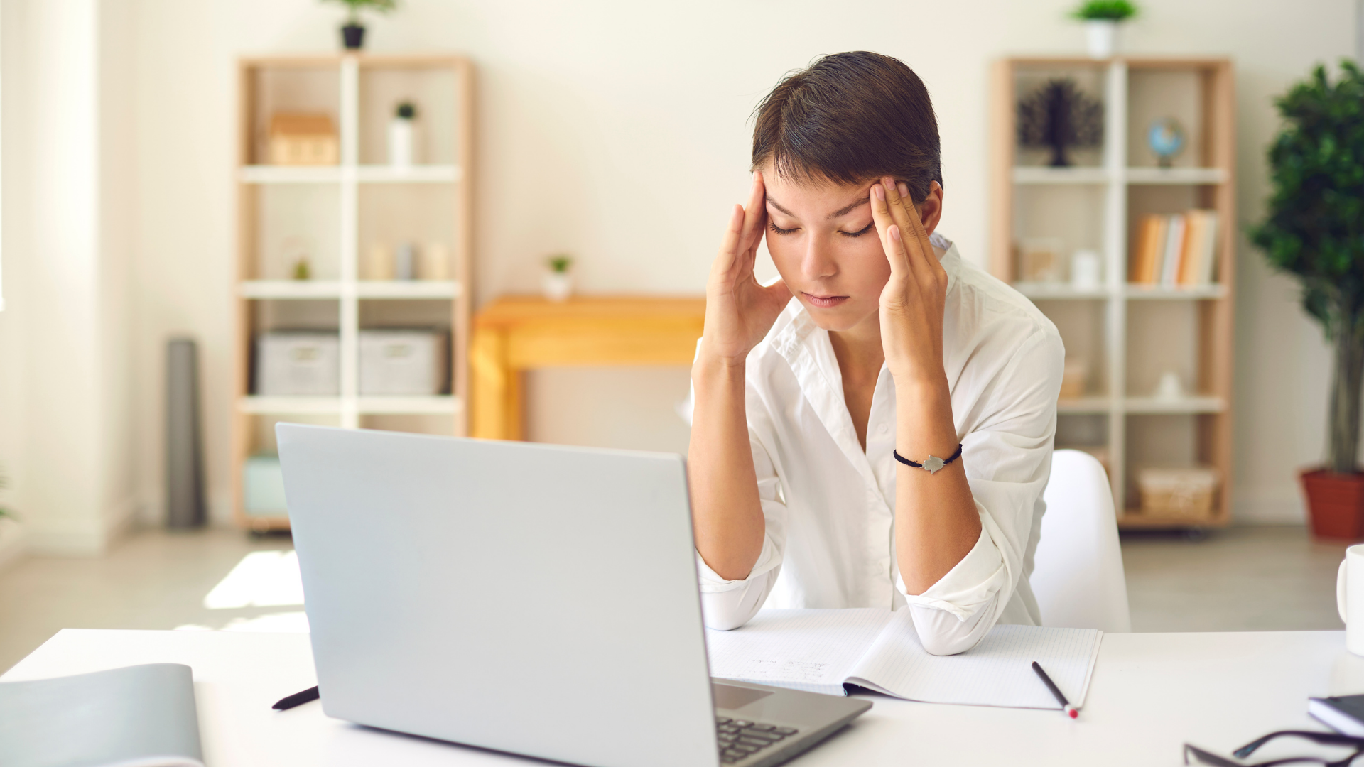 Woman with short hair, stressed, holding her temples while working on a laptop at a desk in a light-filled office.