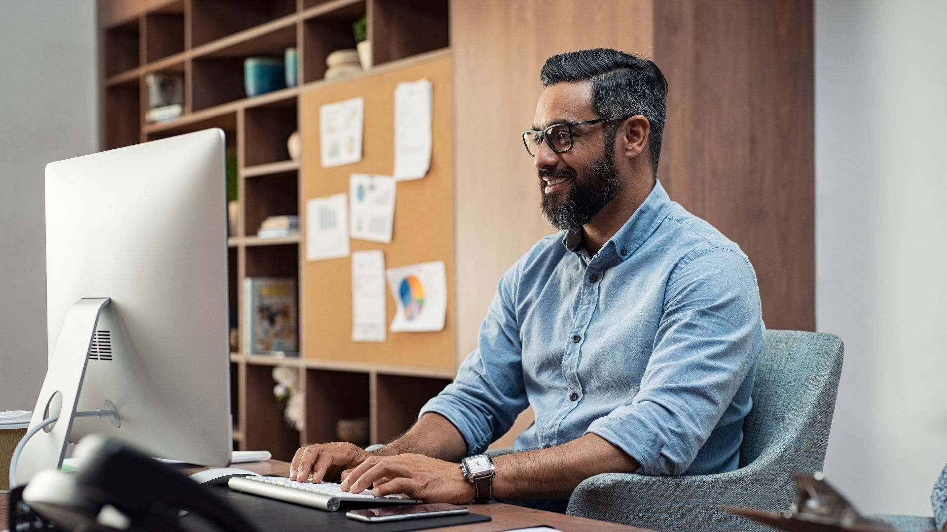 Man with a beard wearing glasses smiles while typing at a computer in a home office setting.