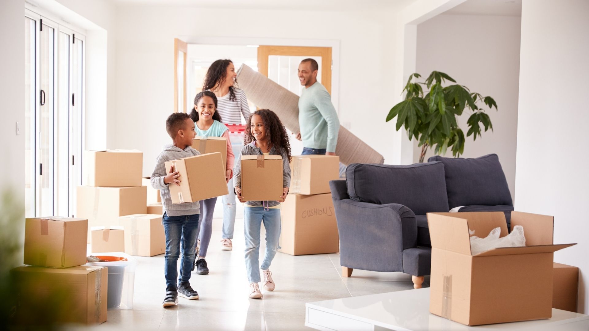 Family moving into a new home, carrying boxes and a rolled-up carpet. Bright, open space with cardboard boxes.