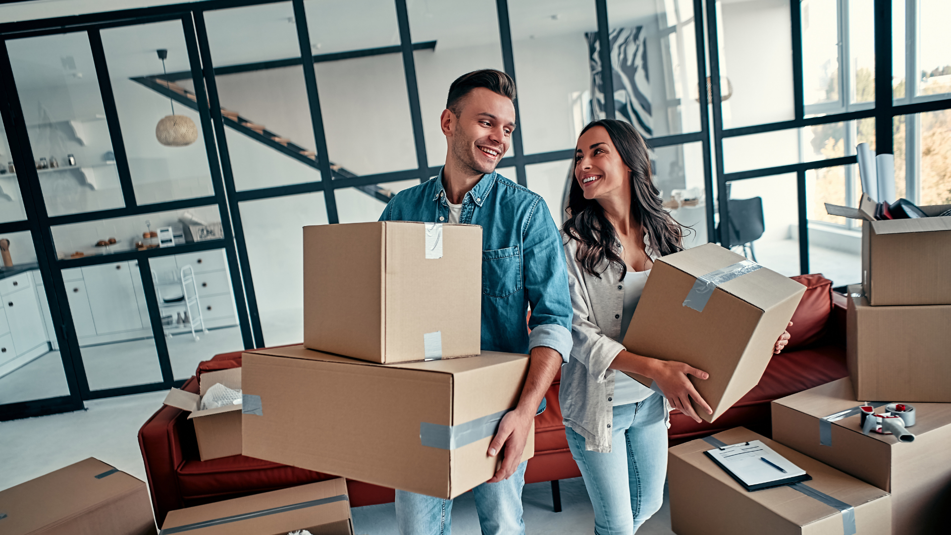 A smiling couple carrying moving boxes in a modern living room with a red couch and glass wall.
