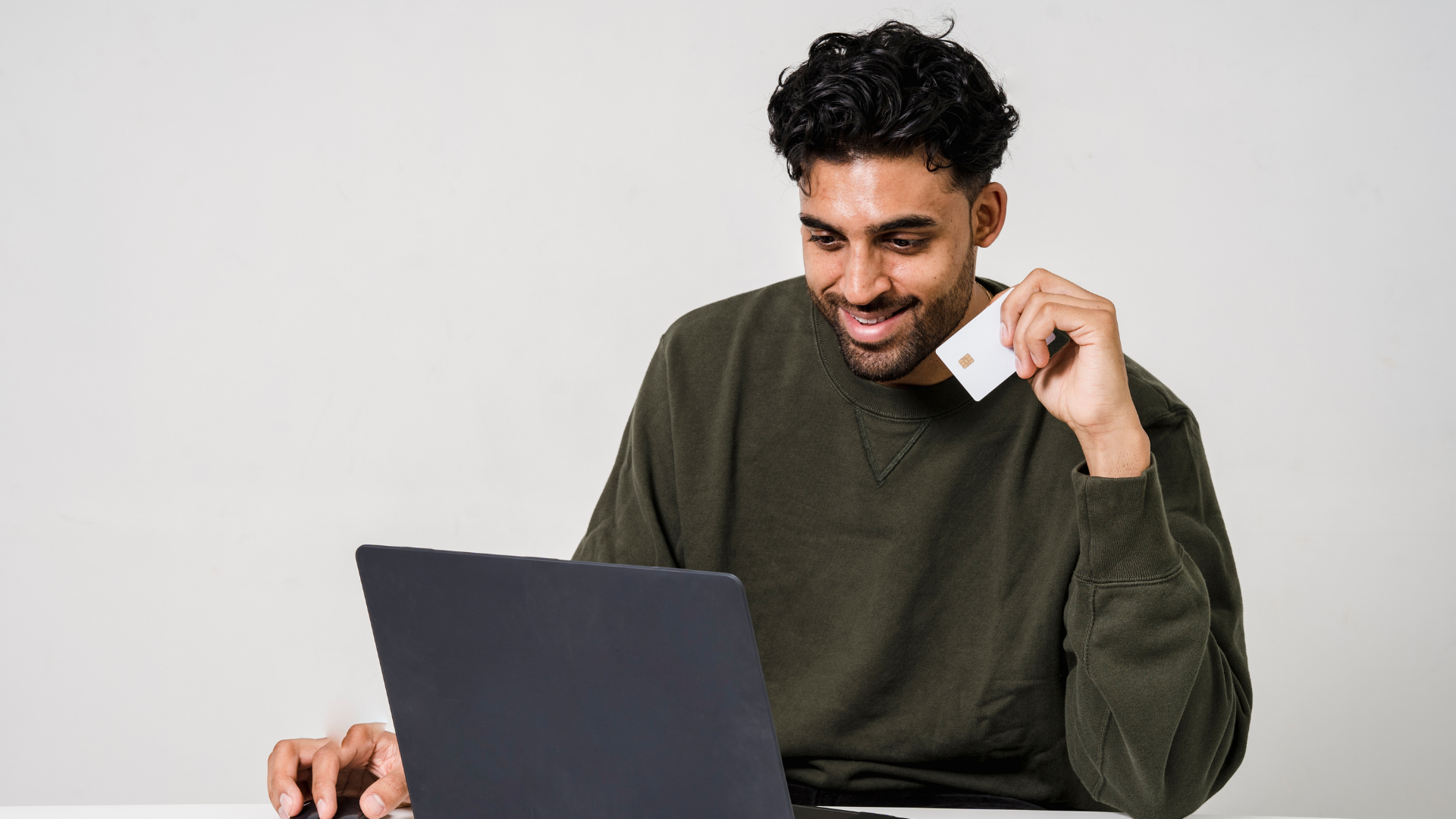 A man with dark curly hair and a green sweatshirt smiles while holding a credit card and looking at a laptop.