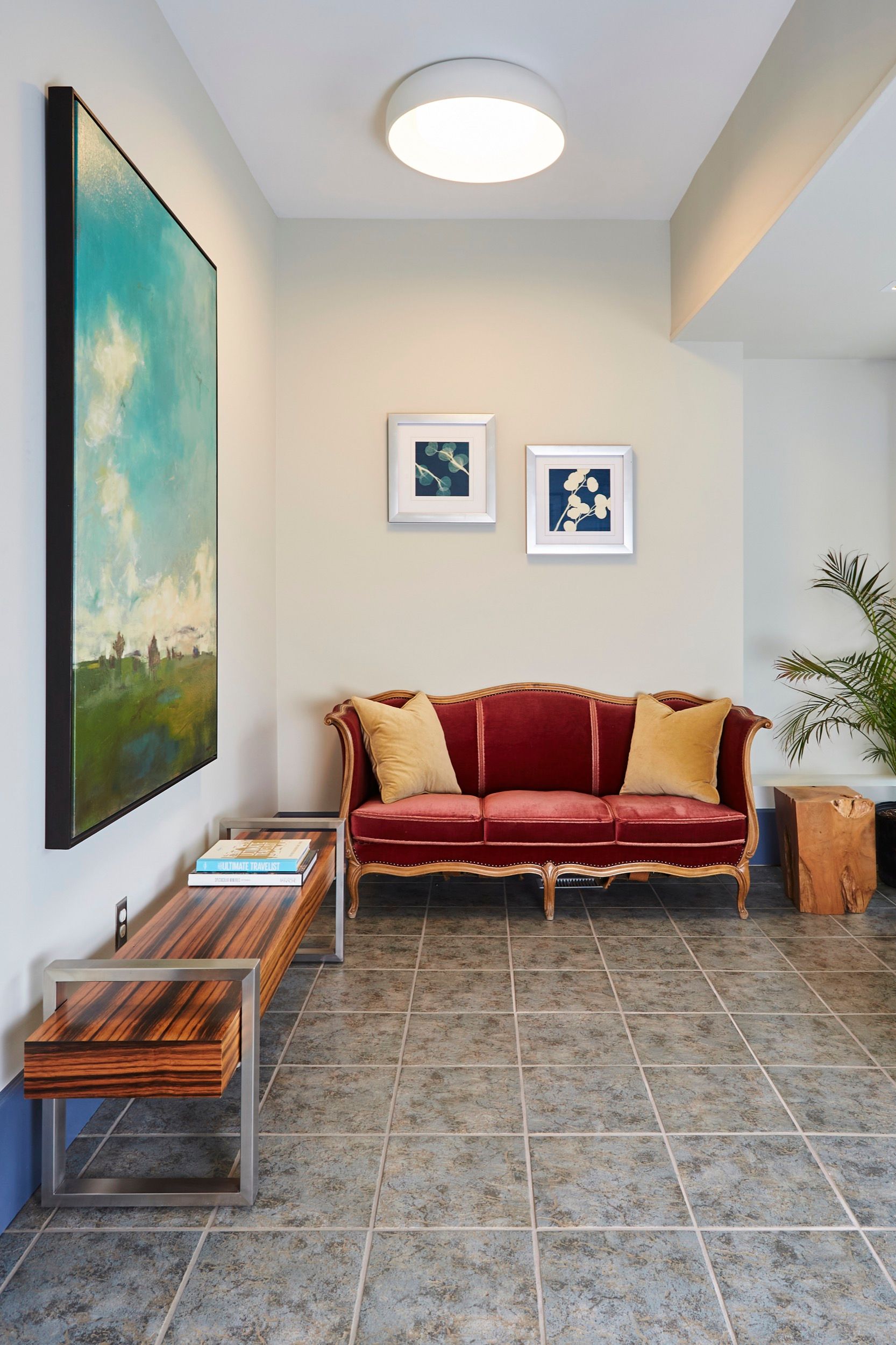A red velvet couch with gold trim, artwork, and a wooden bench in a room with gray tiled floors.