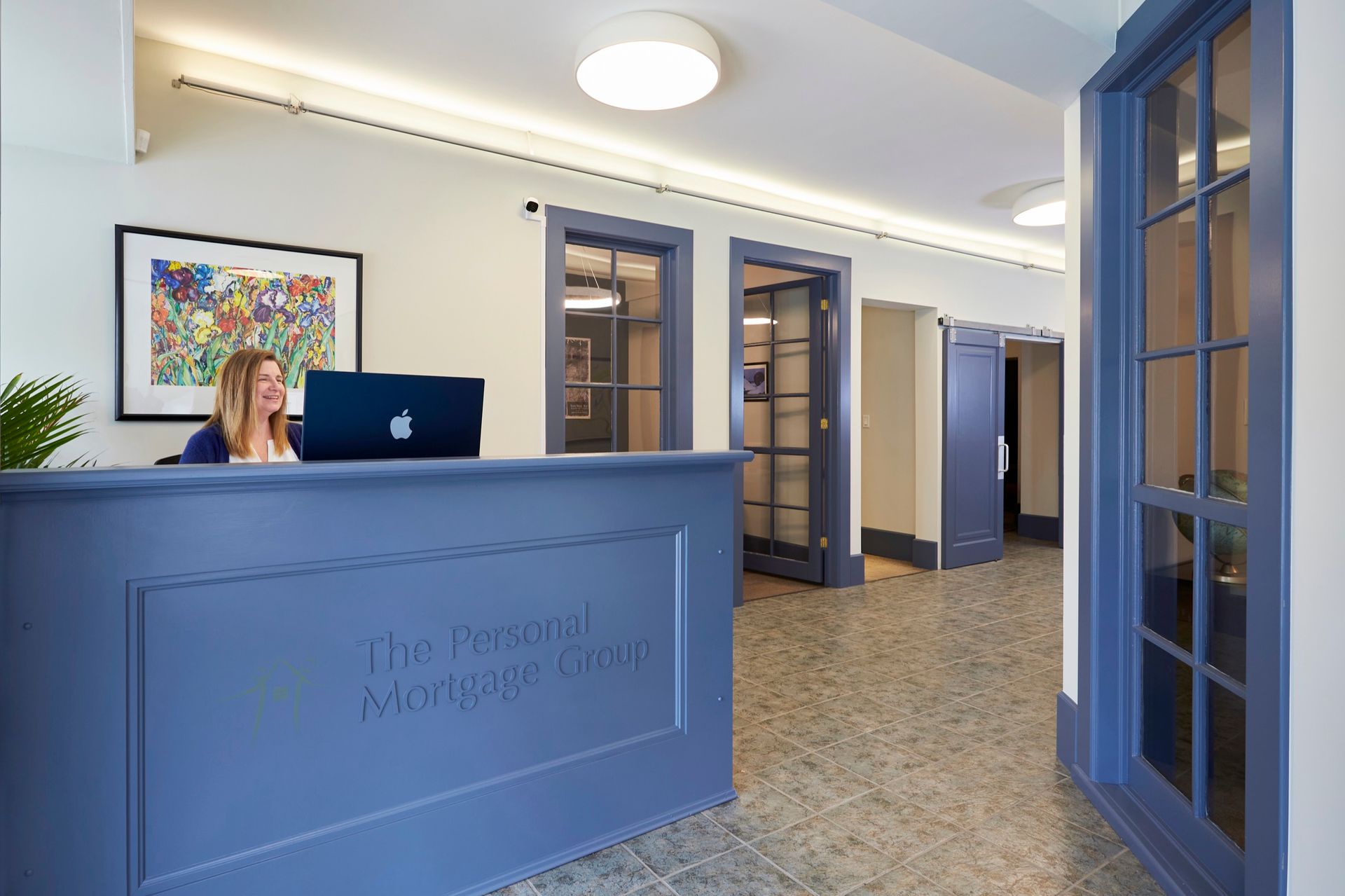 A woman works at a blue reception desk in a well-lit office hallway. Blue doors and artwork decorate the space.