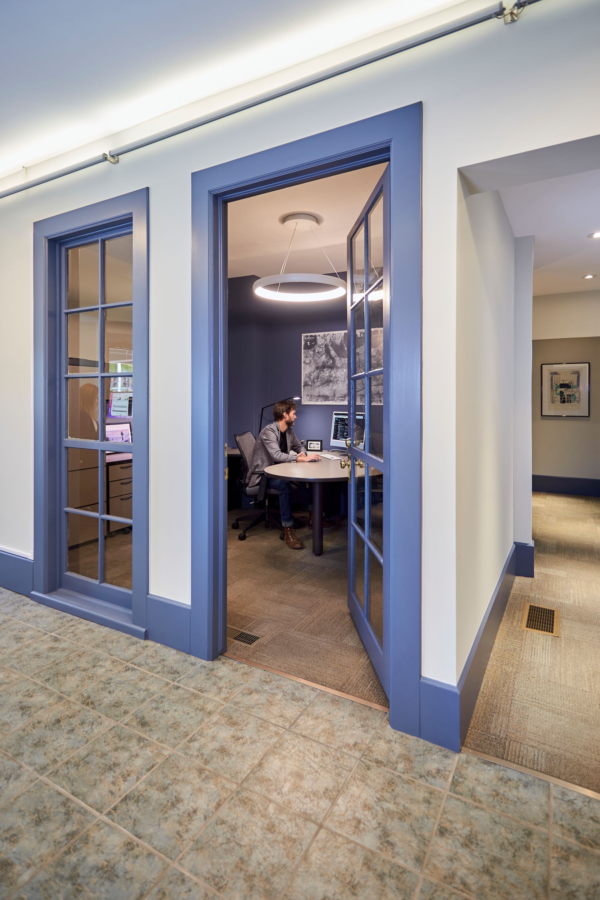 A person works at a desk inside an office with blue-trimmed doors and a neutral-toned hallway.