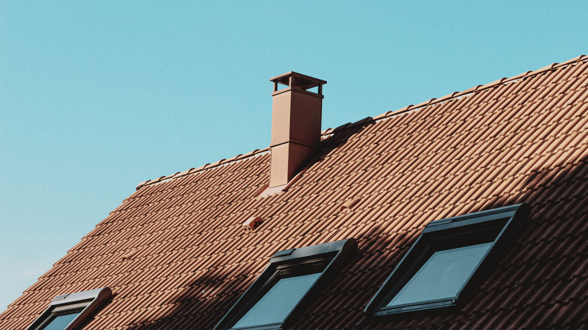 Red tiled roof with chimney and skylights against a clear blue sky.
