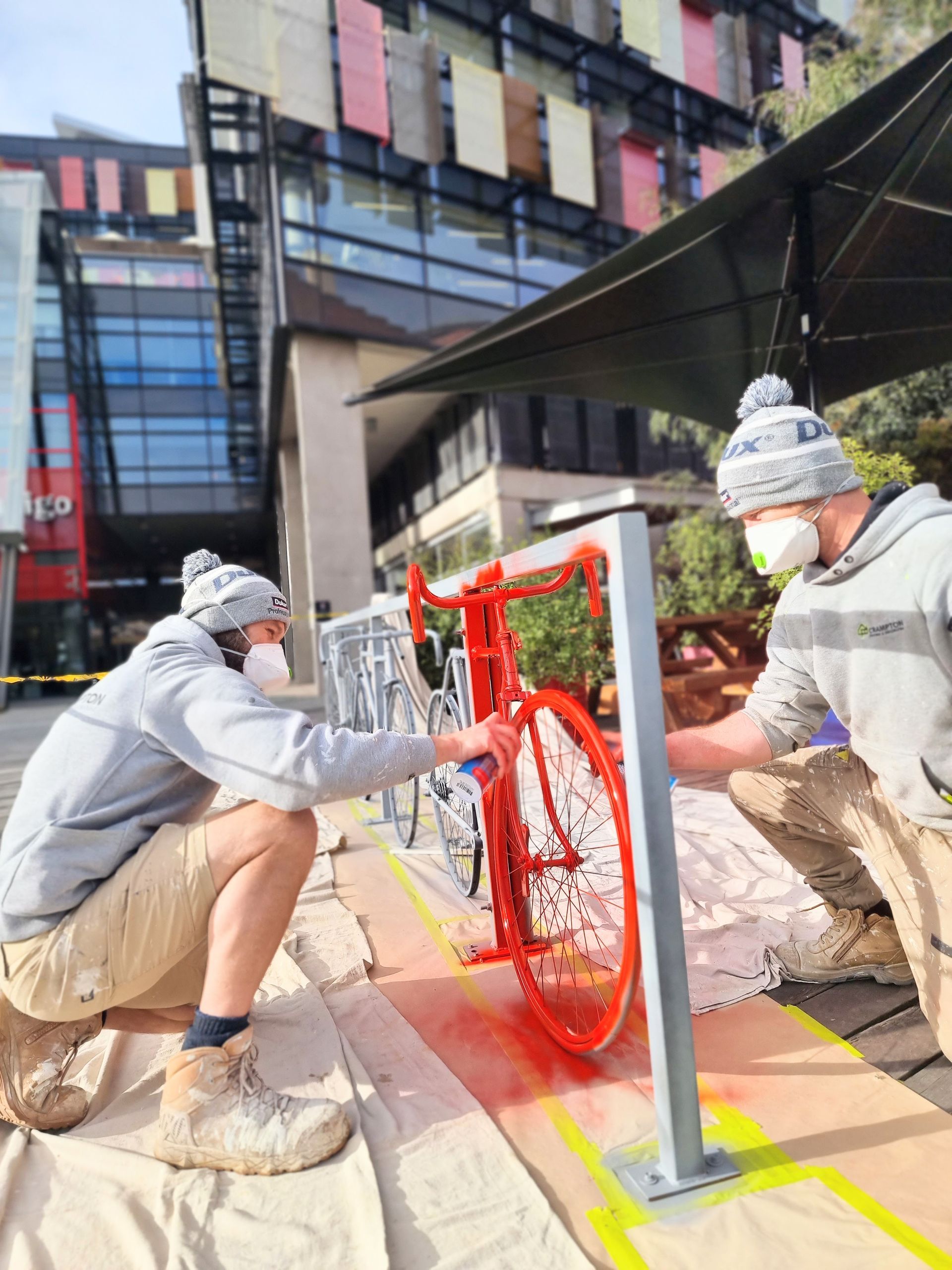 Two people painting a red bicycle at a bike rack outdoors, both wearing masks, beanies, and work clothes — Crampton Painting & Decorating Pty Ltd In Maiden Gully, VIC