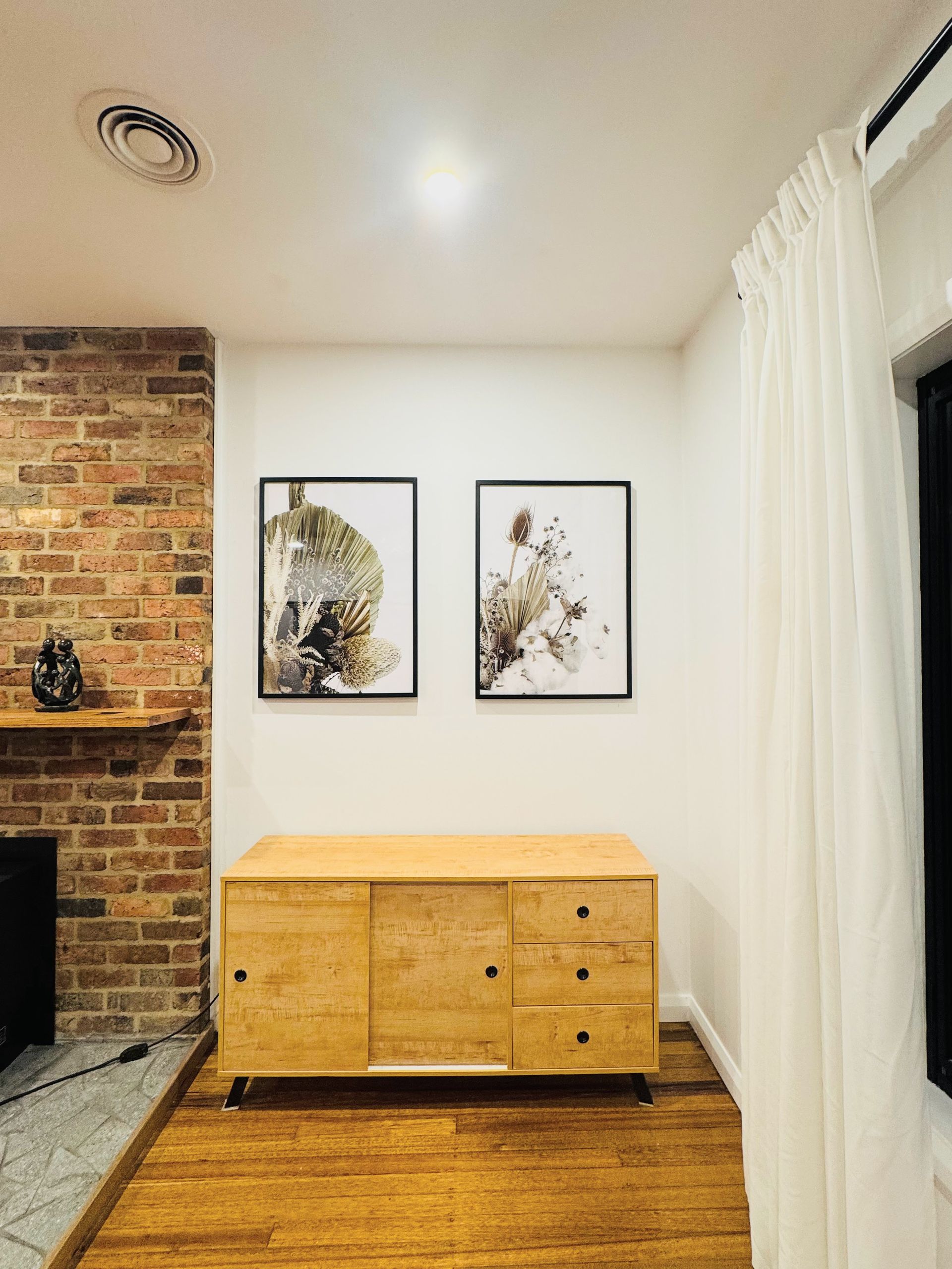 Wooden cabinet with two framed art pieces on white wall. Brick fireplace to the left, curtained window on right — Crampton Painting & Decorating Pty Ltd In Echuca, VIC