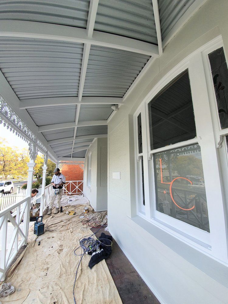 A Man Is Standing On A Porch Painting A House — Crampton Painting & Decorating Pty Ltd In Maiden Gully, VIC