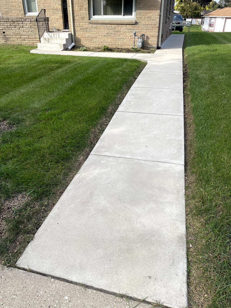 A concrete walkway leading to a house next to a lush green lawn