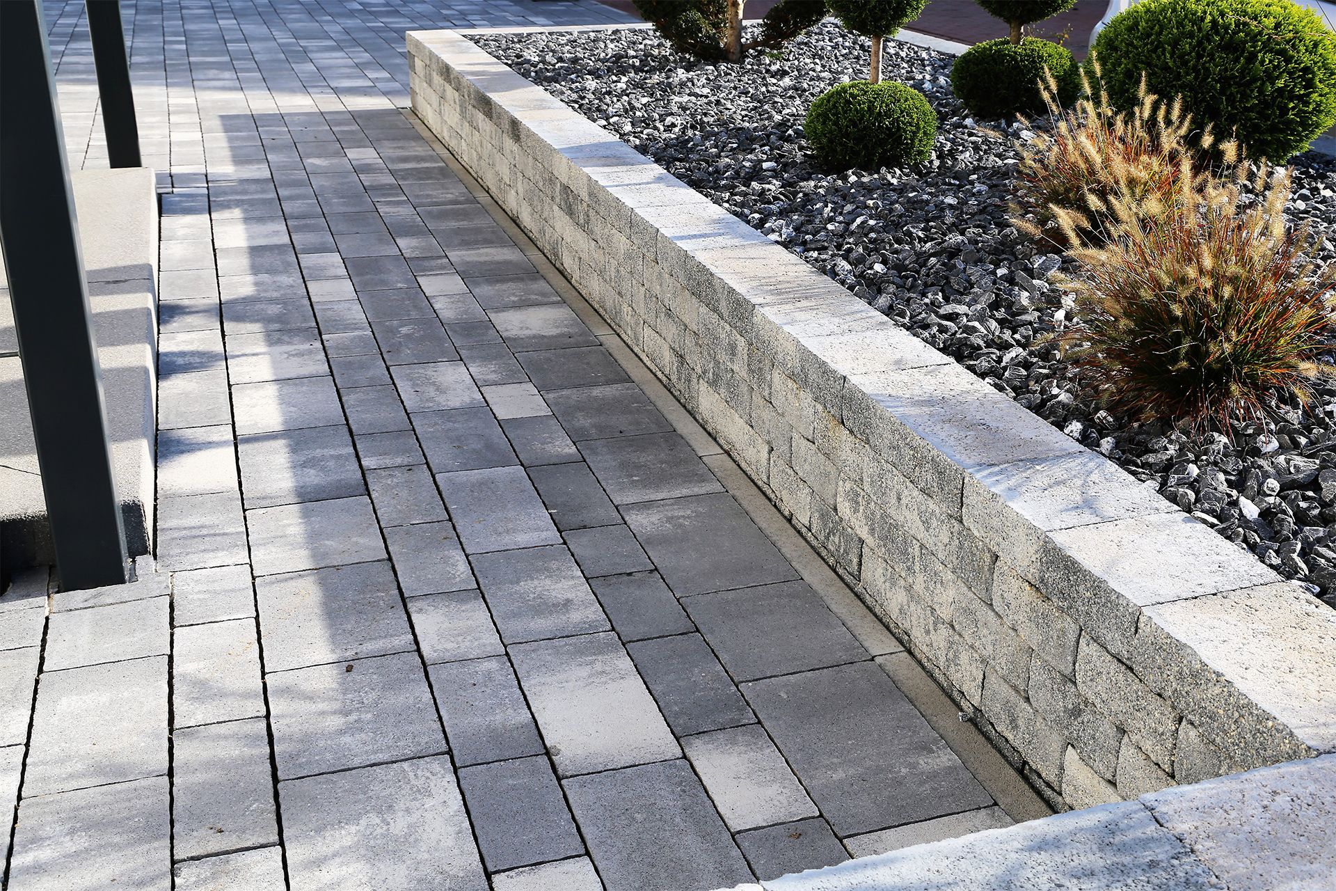 Pathway and retaining wall made of gray paving stones, with shrubs and dark gravel in a garden.