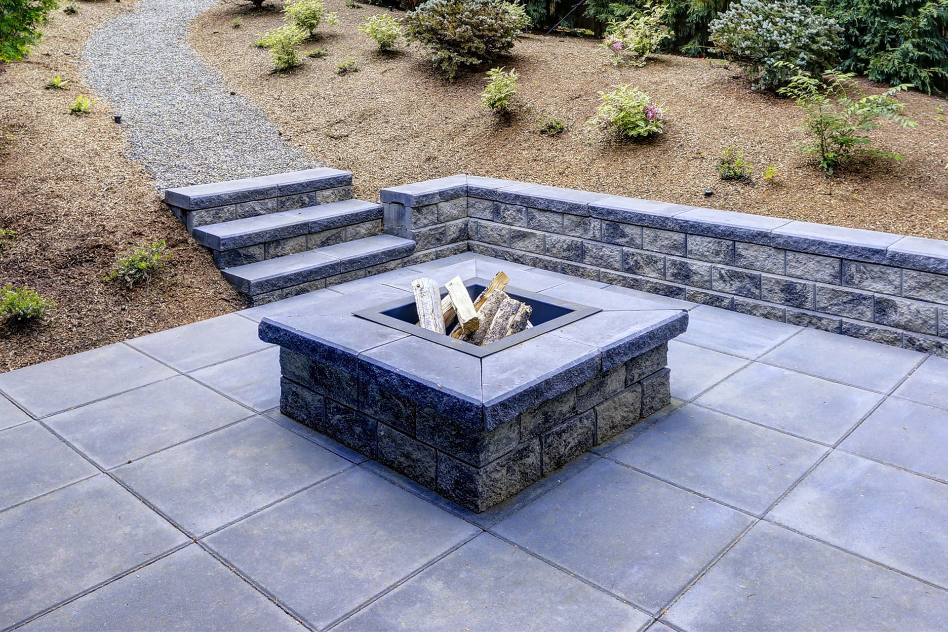 Patio with stone fire pit, retaining wall, steps, and gravel path on a hillside.