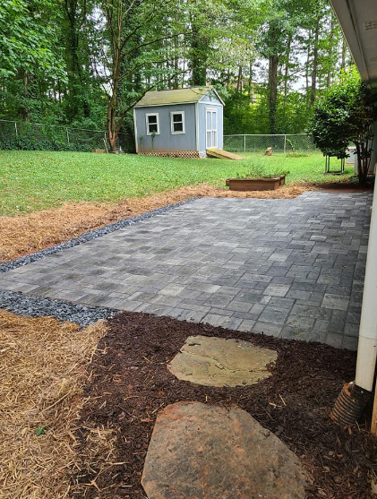 Newly constructed gray paver patio with mulch and stepping stones. Shed and green yard in background.