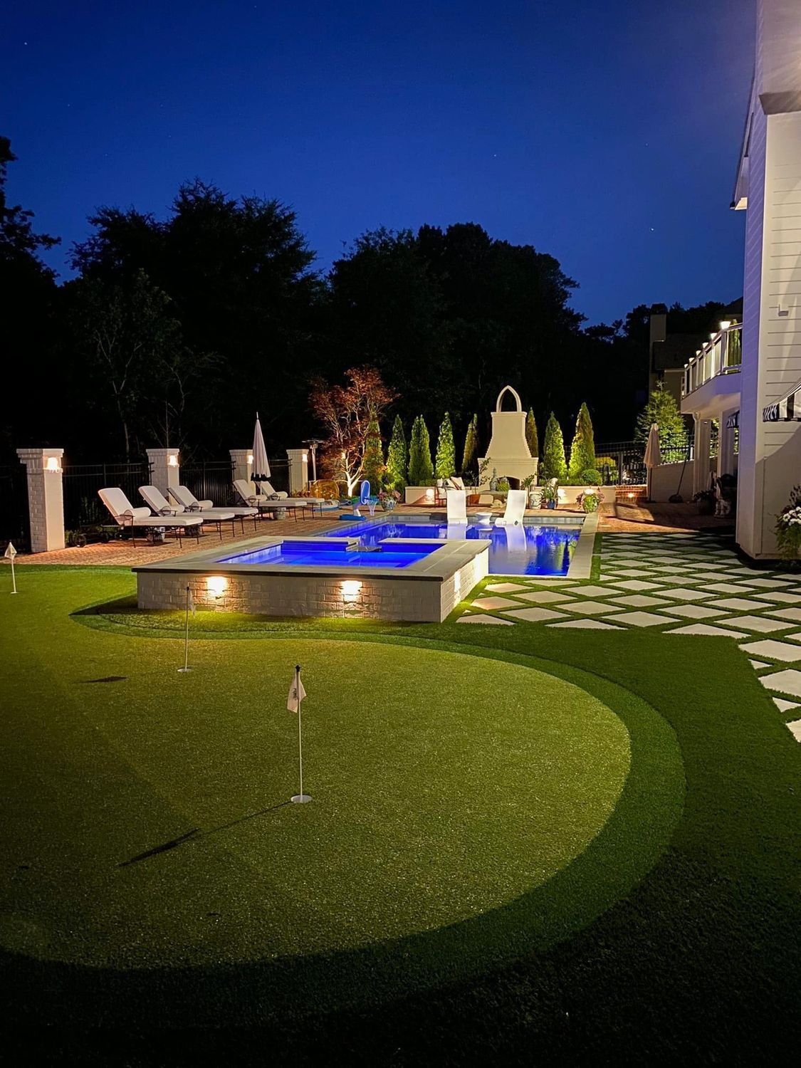 Night view of a luxury backyard with a pool, putting green, lounge chairs, and architectural features.