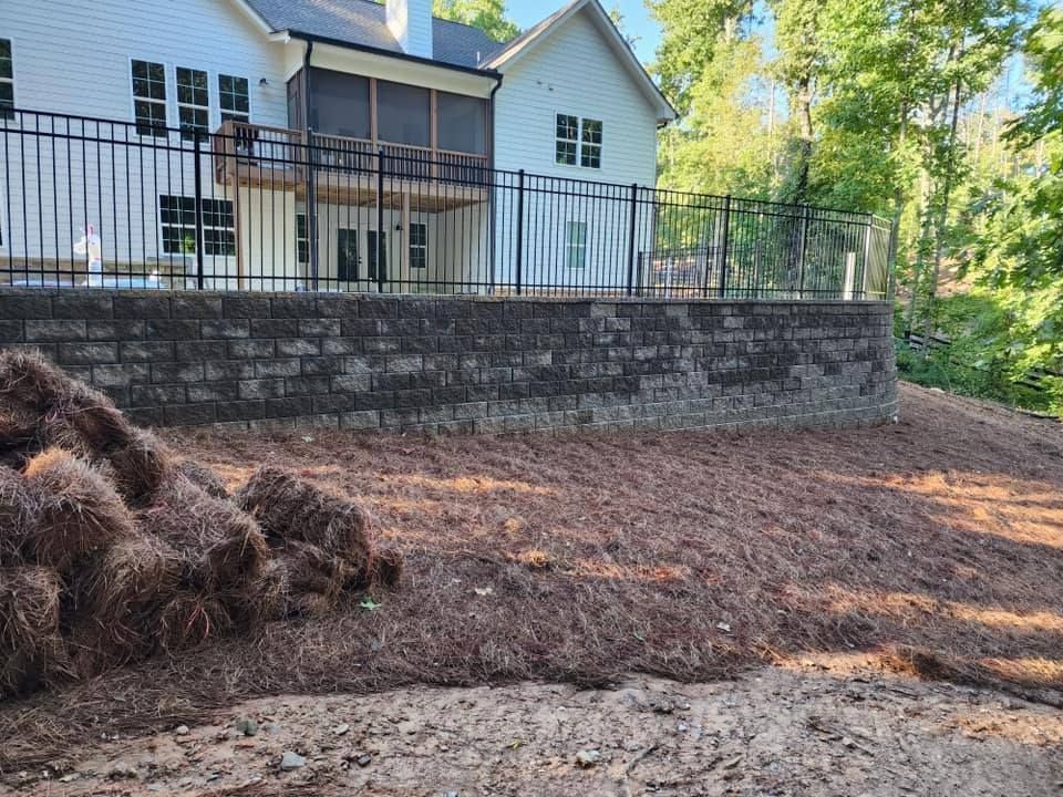 Retaining wall in front of a white house with black fence, pine straw ground cover.
