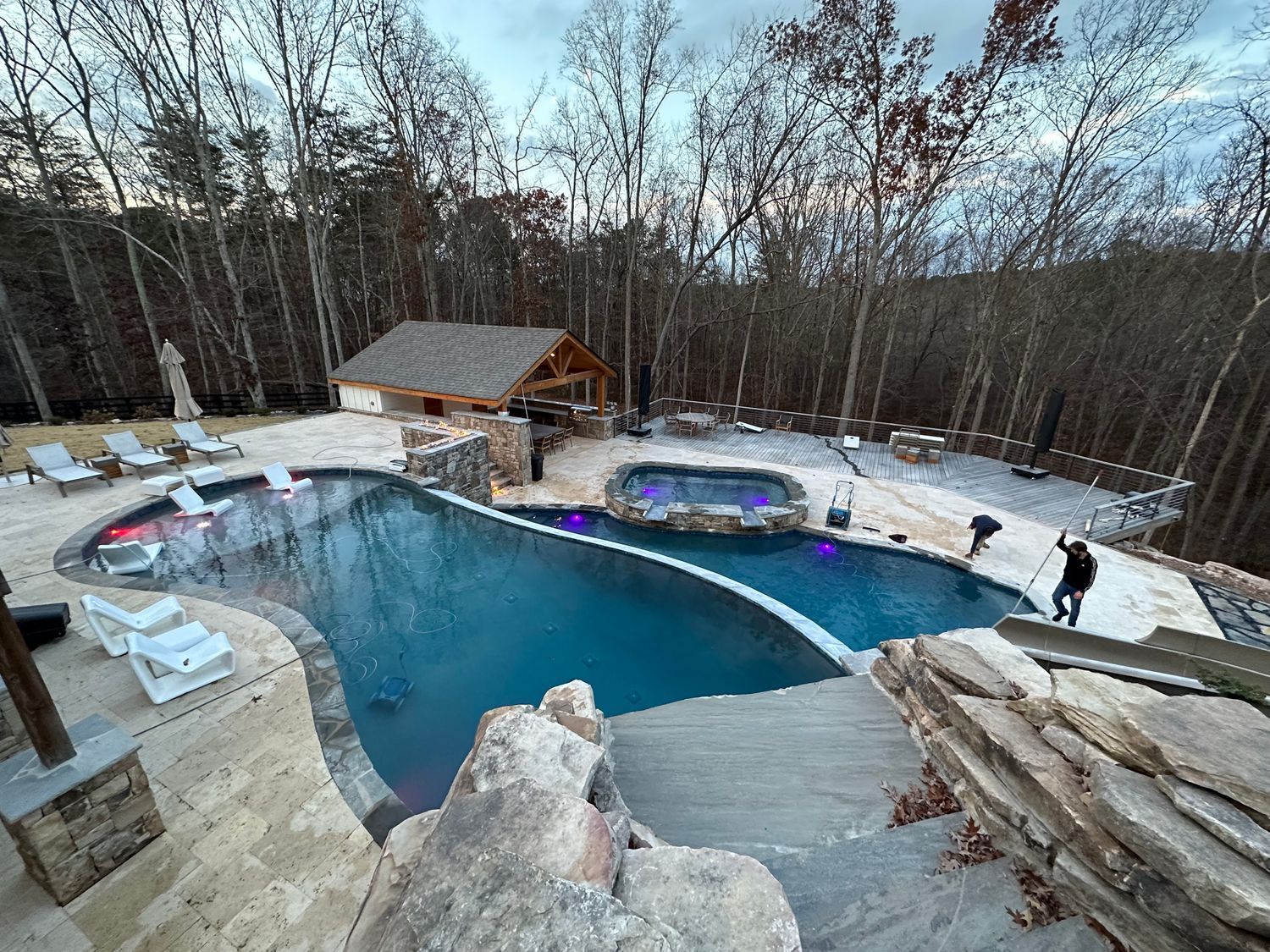 Pool and hot tub surrounded by trees, stone features, and a small cabana.