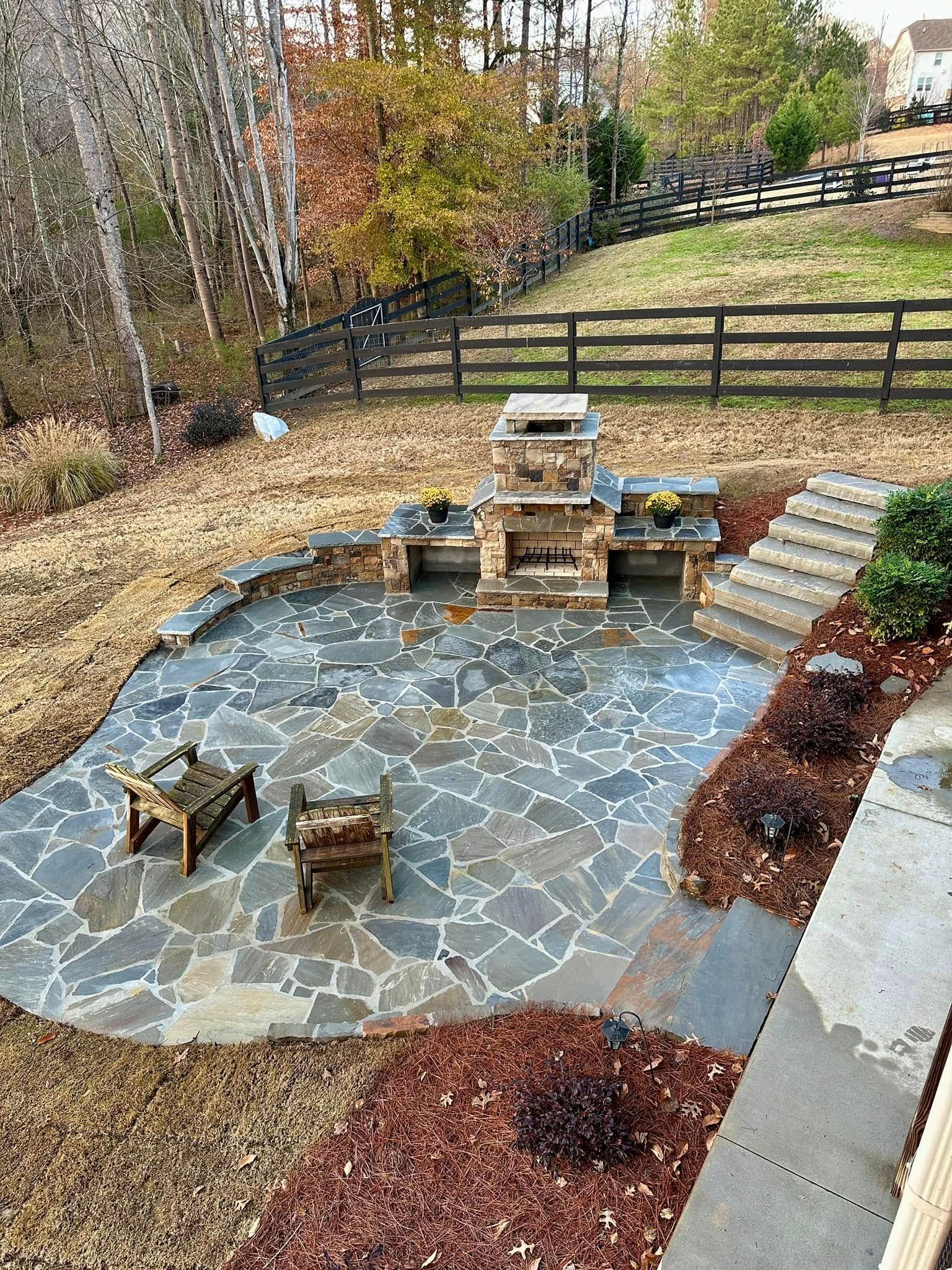 Patio with stone fireplace and chairs, surrounded by landscaping and fence.