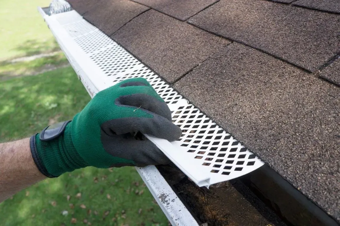 A gloved hand installing a white, perforated plastic gutter guard over a dark shingled roof.