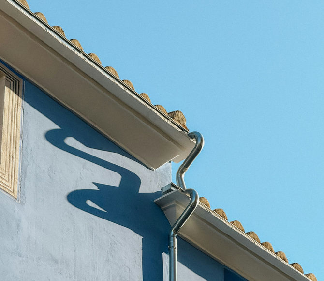 A metallic gutter system on the corner of a light blue building, casting a unique, abstract shadow on the wall.