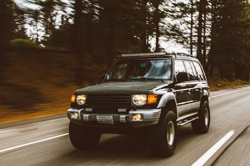 Black SUV driving on a road through a forest, amber headlights on.