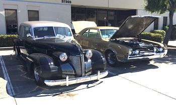 Two old cars are parked in a parking lot with their hoods up.