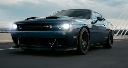 Dark blue Dodge Challenger driving on a road. The car has black wheels and a prominent hood scoop; bright headlights are on.