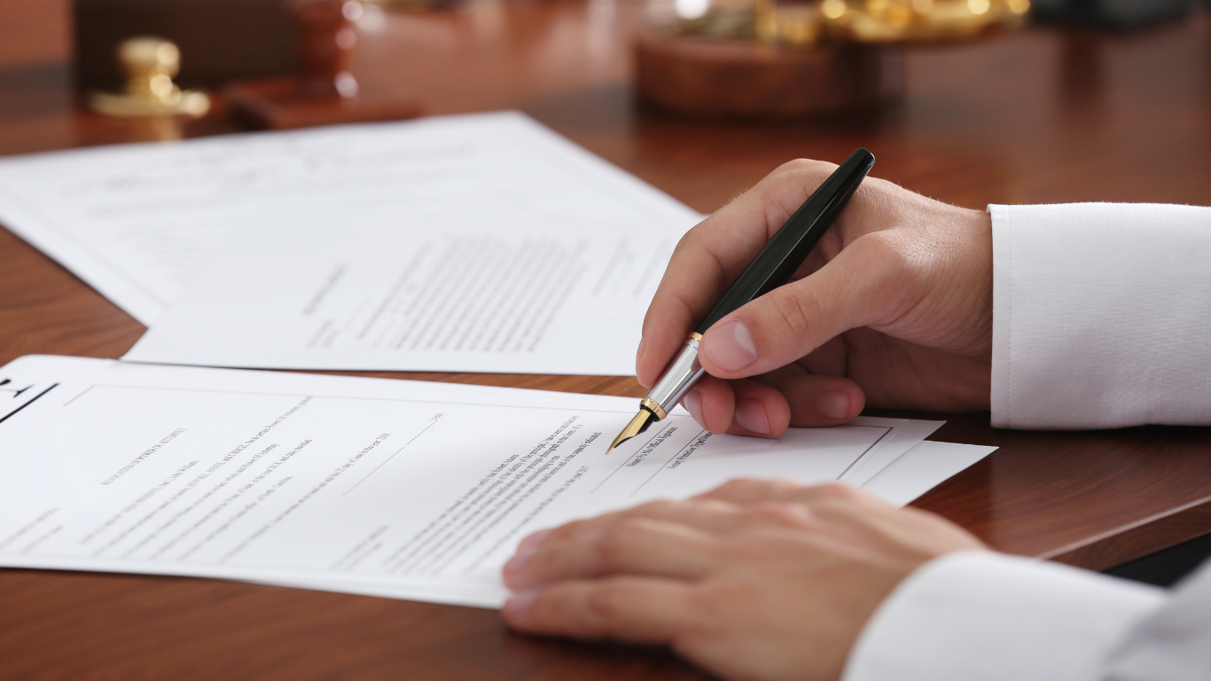 Person signing legal documents with a pen on a wooden desk.