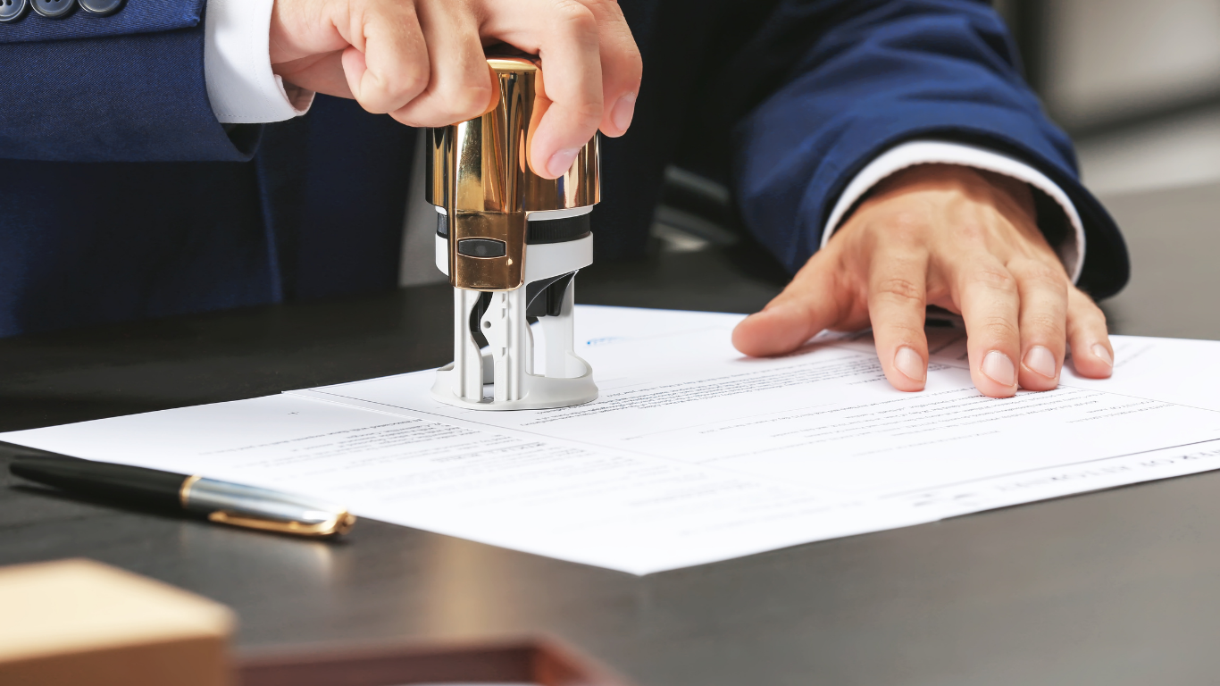 Person in blue suit stamping a document with a golden stamp. A pen and small box are also visible.