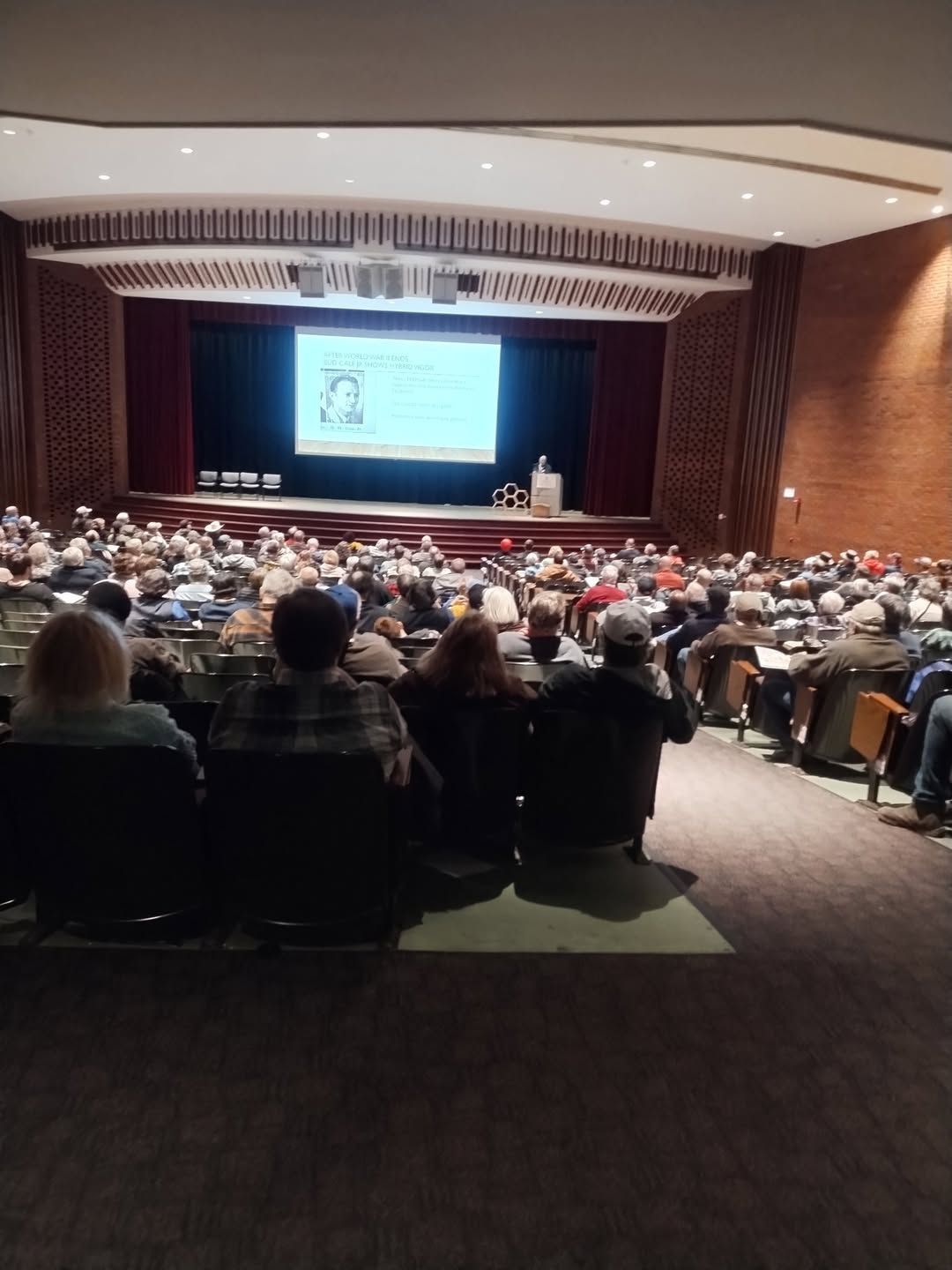 A large group of people are sitting in an auditorium watching a presentation on honeybees.