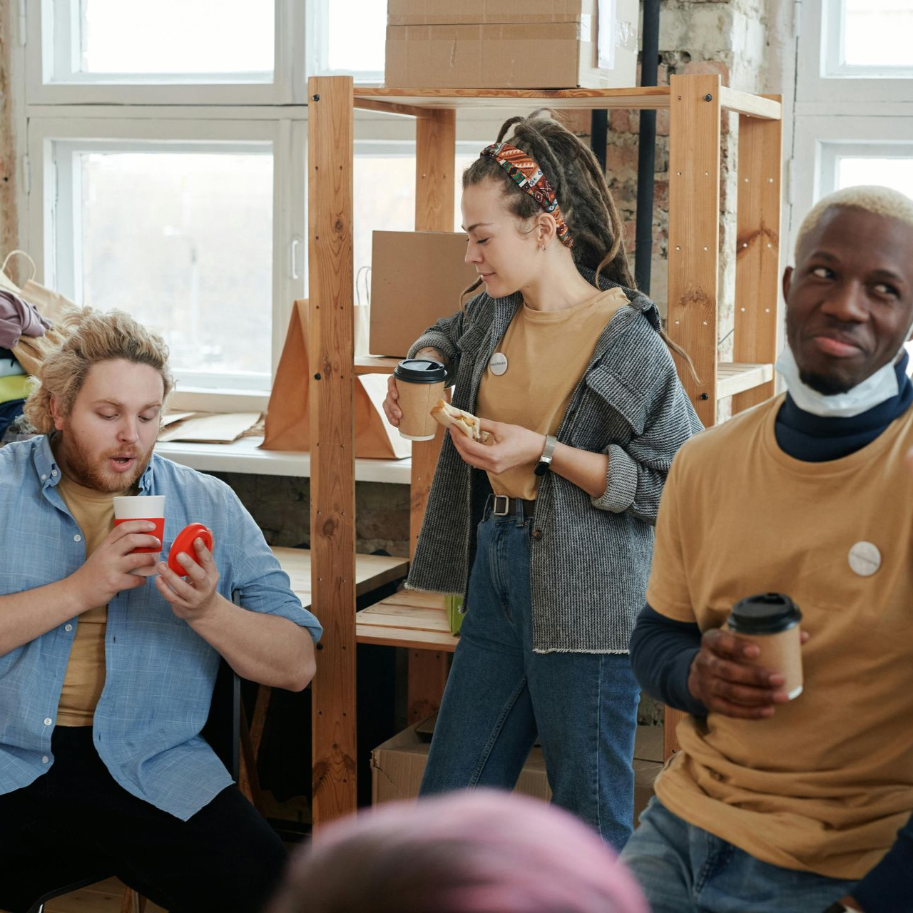 Dans un studio lumineux, trois personnes discutent en tenant des tasses de café près d'une grande fenêtre.