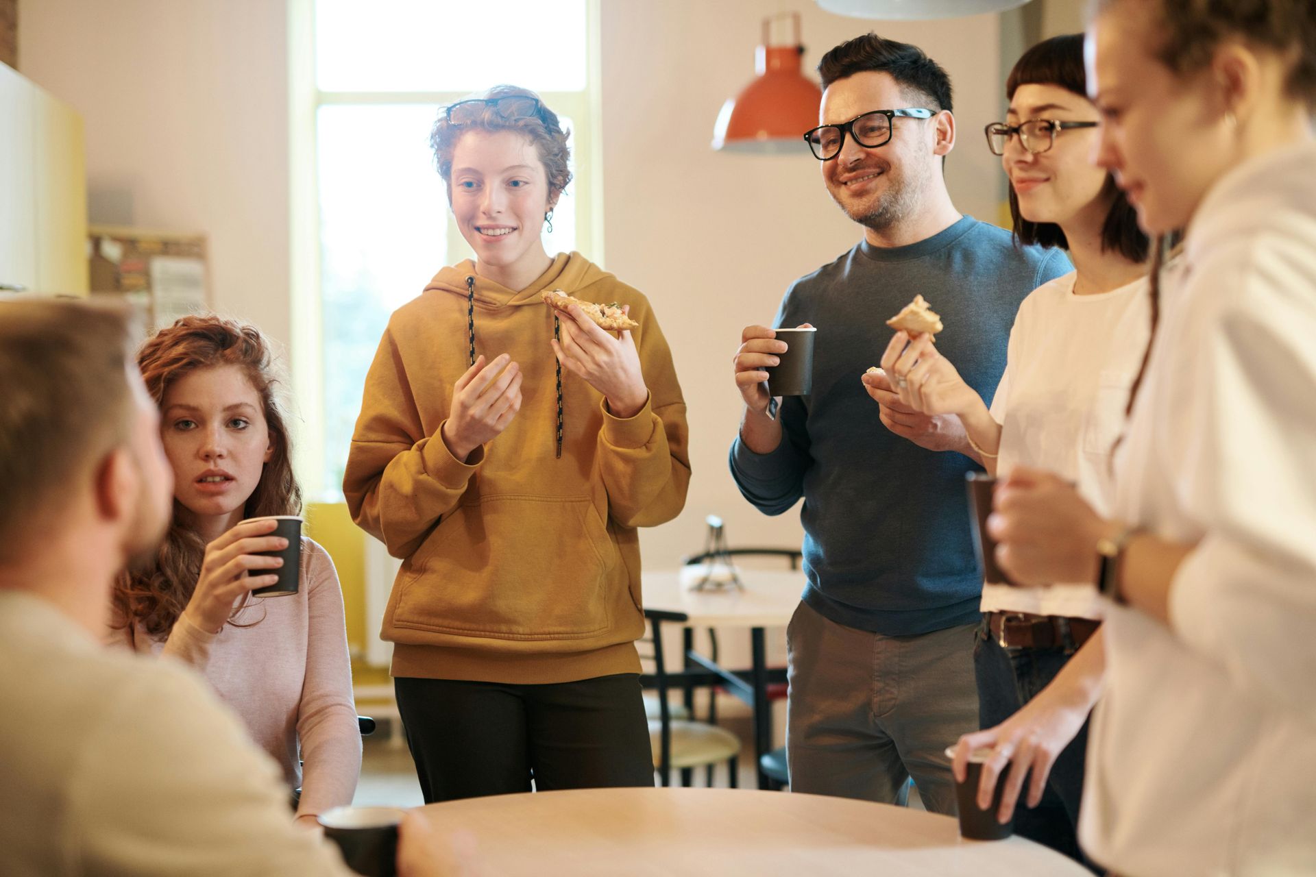 Friends standing in a bright kitchen, eating snacks and chatting around a table