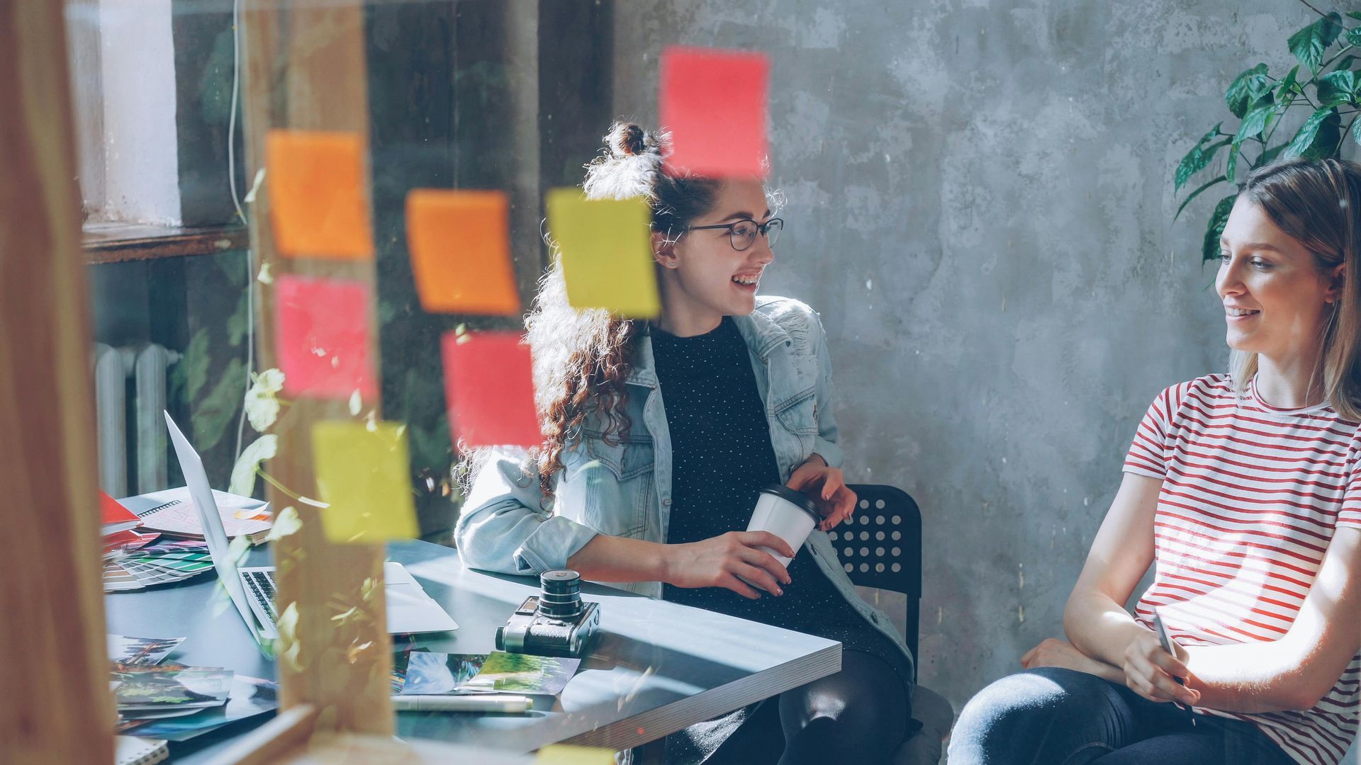 Two people chatting in an office, with colorful sticky notes on a glass wall.