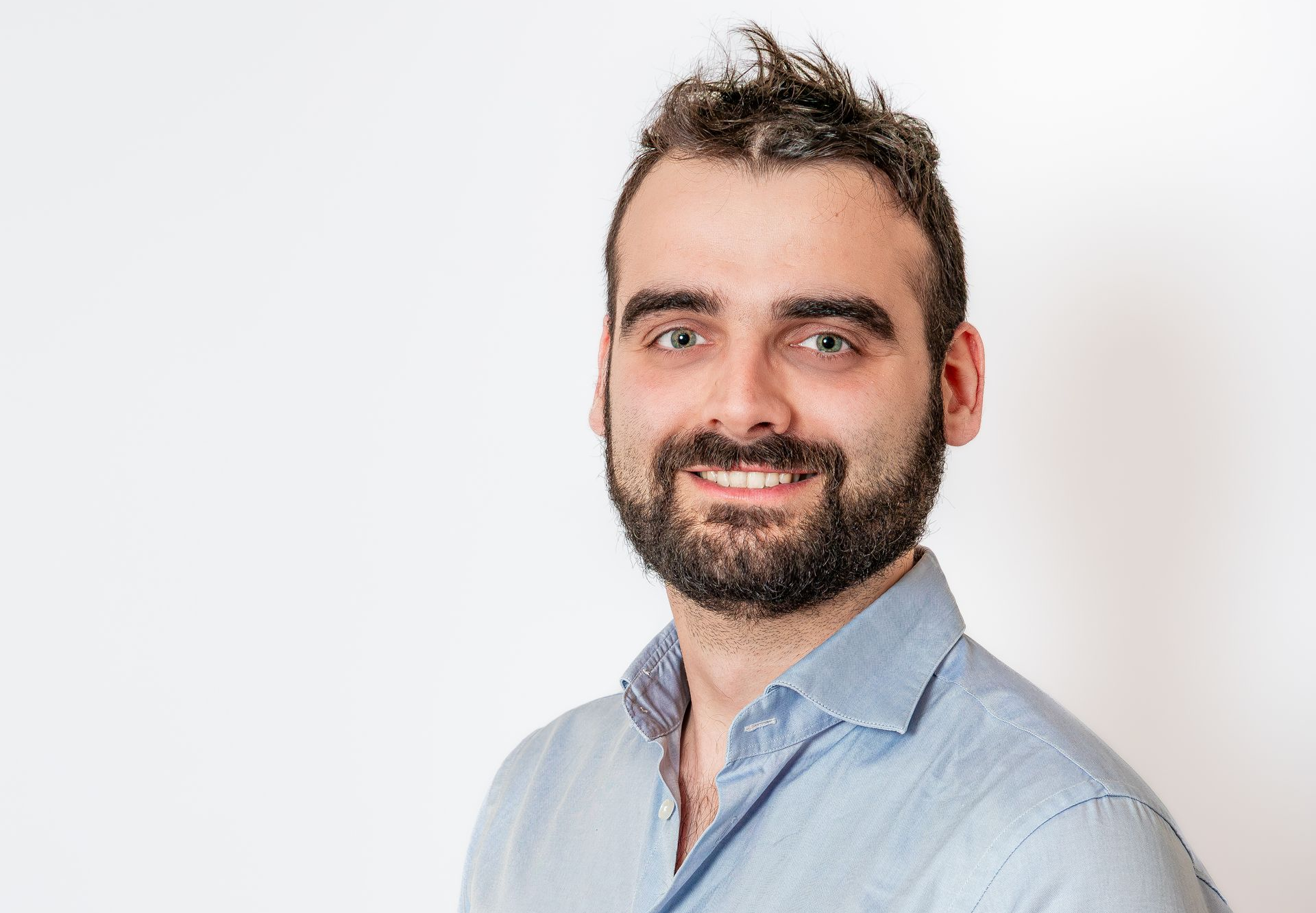 Homme souriant aux cheveux courts et foncés et à la barbe, portant une chemise bleu clair sur fond blanc