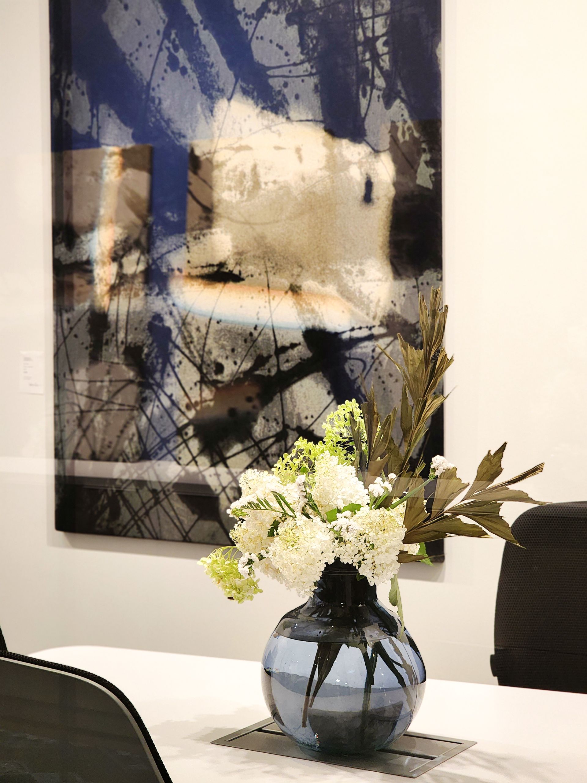 A vase of flowers is on a table in front of a painting.