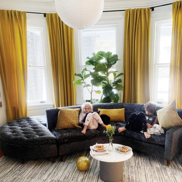 Two children sit on a couch in a living room with yellow curtains