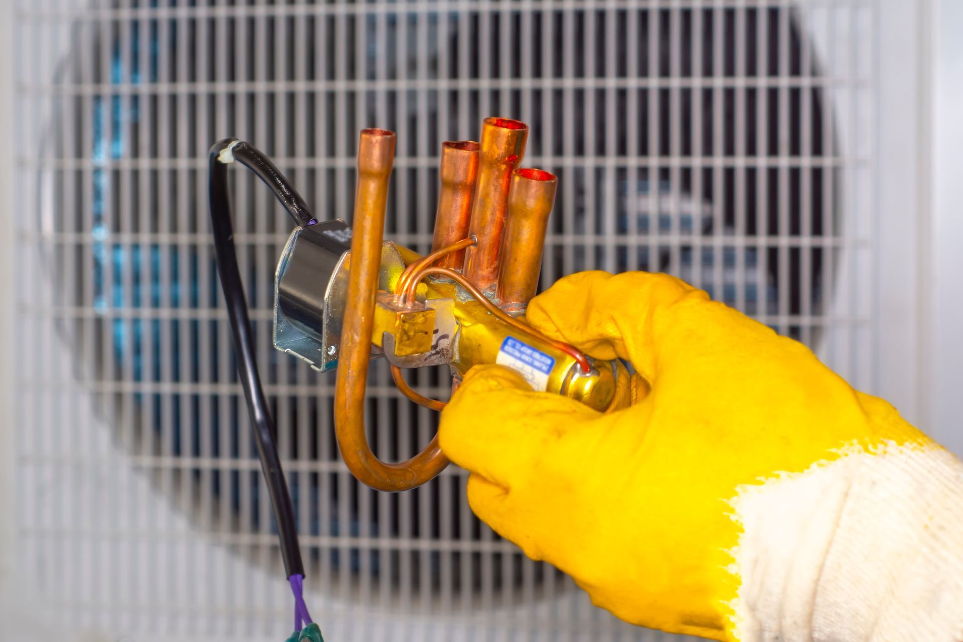 A person wearing yellow gloves is holding a copper pipe in front of an air conditioner.