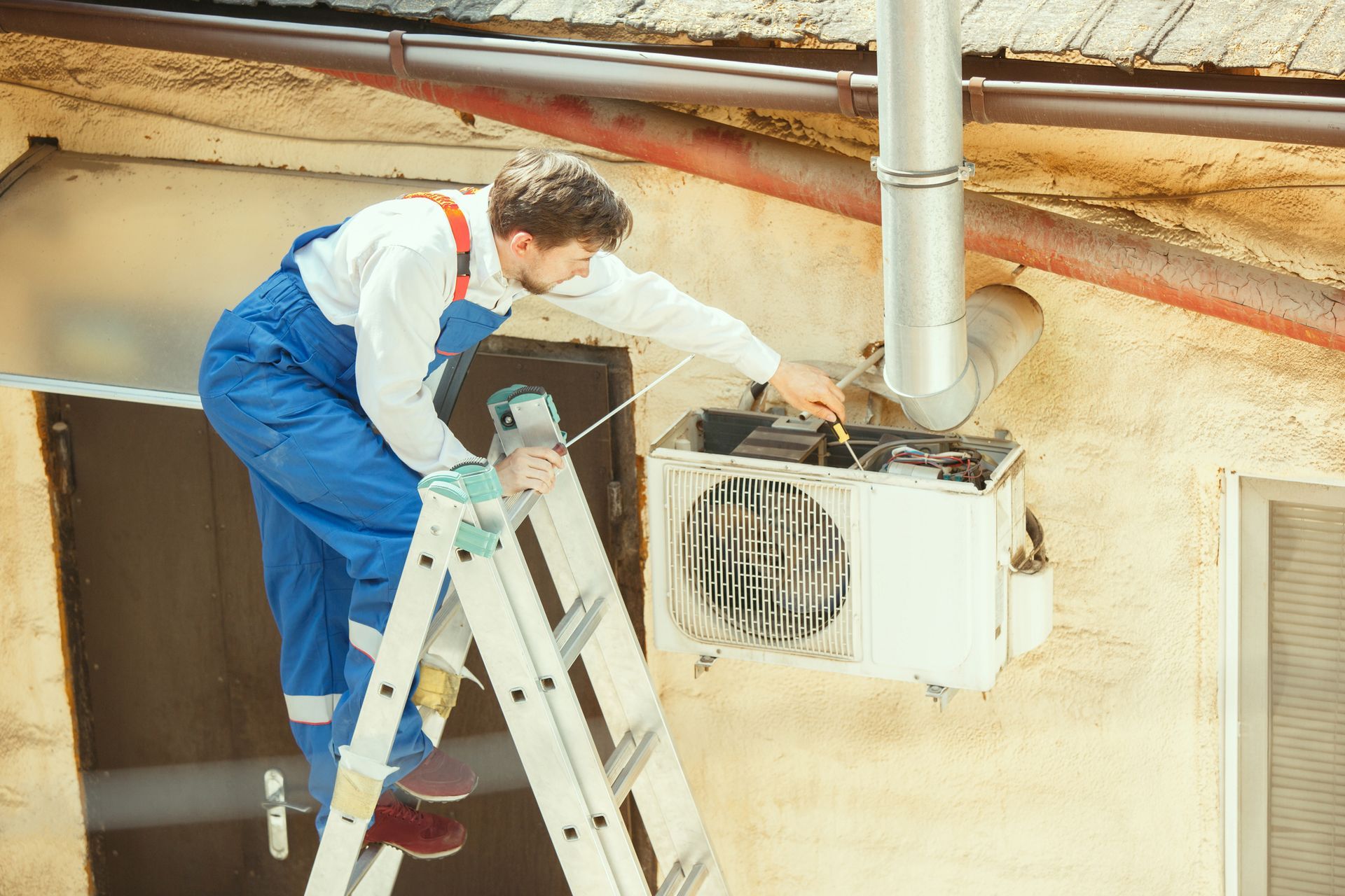 A man is sitting on a ladder fixing an air conditioner.