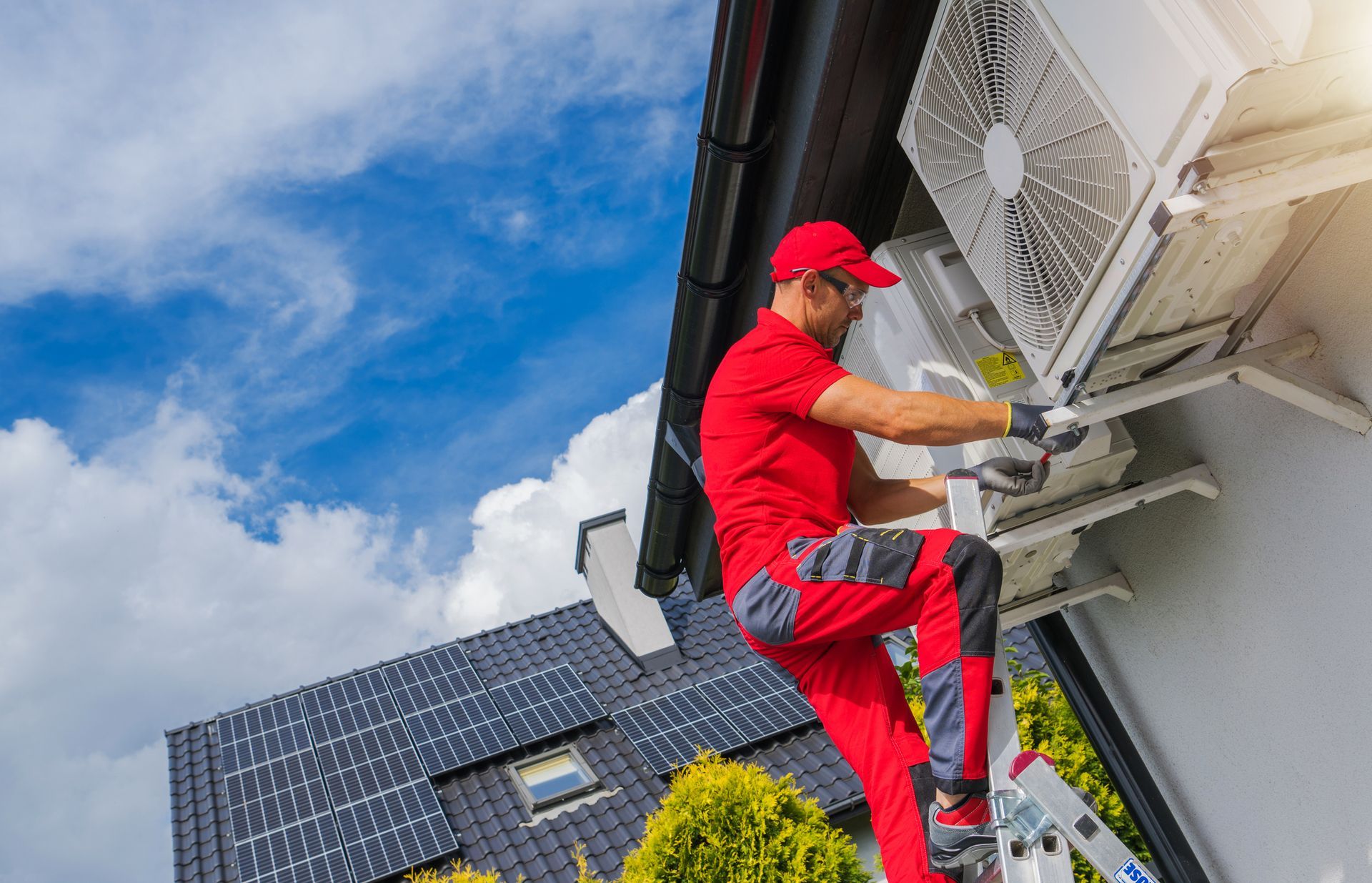 A man is sitting on a ladder fixing an air conditioner on the side of a building.