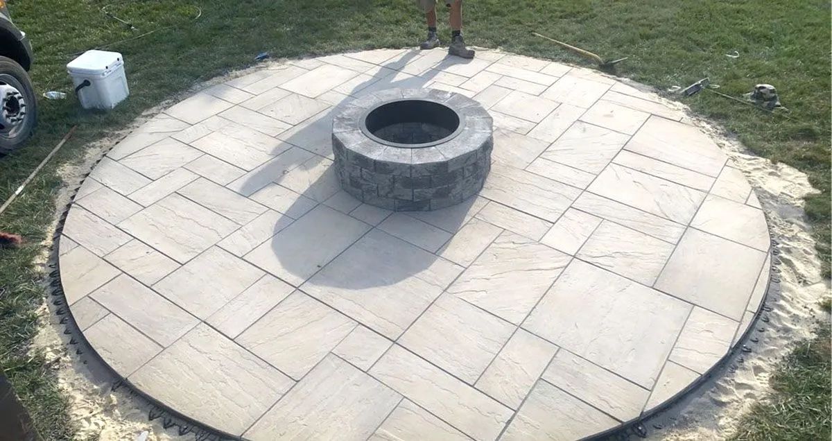 Circular stone patio with a round fire pit in the center on a grassy lawn.